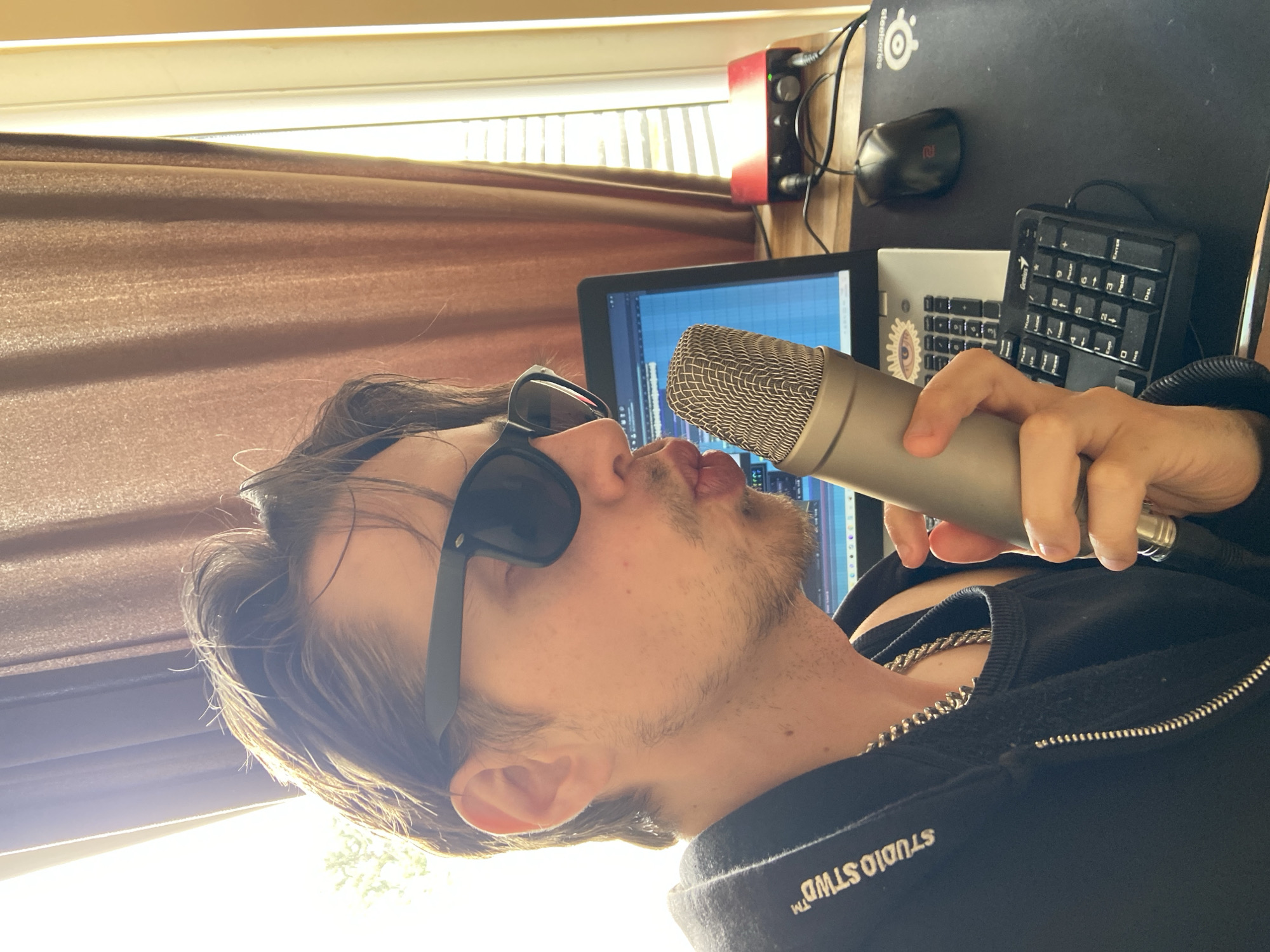 A man wearing sunglasses is holding a silver microphone to his mouth. He is wearing a black hoodie with a silver chain around his neck. Behind him is a computer monitor displaying audio editing software, a keyboard, a number pad, a computer mouse, and an audio interface. A window with brown curtains is visible in the background. The location is not discernible from the image.
