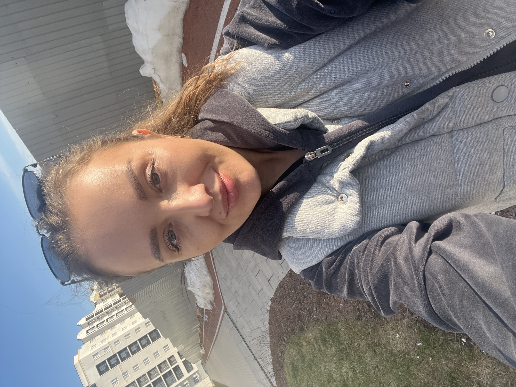 A selfie taken outdoors, showing a young woman wearing a gray and black hooded jacket. The background reveals a cityscape with a tall, white building visible in the lower left and some green grass and pavement. Some white snow is present. Due to the lack of context information, it is impossible to verify that the selfie was taken in the Russian city of Pervomaysky. No actions or interactions are depicted in the photo.