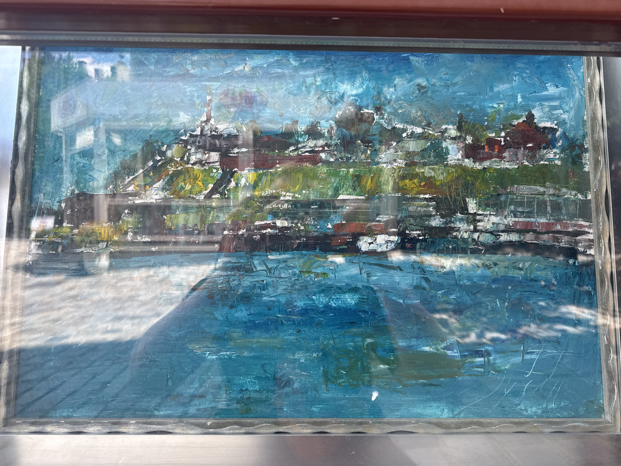 The image shows a framed, impasto oil painting depicting a waterfront scene, likely Kalemegdan Fortress in Belgrade, Serbia. The painting features a dense, layered application of oil paint, creating a textured, abstract representation of buildings and water. Red-brown structures, reminiscent of the fortress walls and buildings, are visible against a bright blue sky and a turquoise and deep blue body of water in the foreground. The water reflects the sky and the buildings, with visible brushstrokes suggesting movement and light. There are no people or animals depicted in the painting. The painting is framed and covered by glass, which reflects the environment, including a shadow and possibly a ceiling. The overall impression is one of a vibrant, somewhat impressionistic, urban landscape painting.