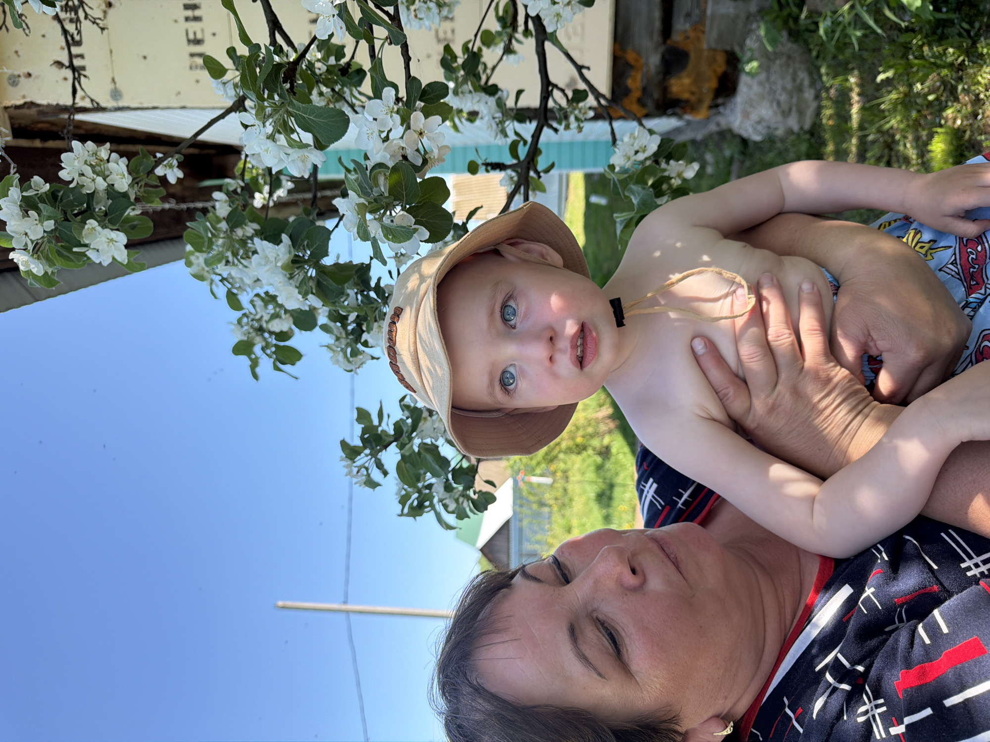 The image features a child being held by an adult, likely outdoors. The child has fair skin, blue eyes, and is wearing a tan sun hat. The adult, presumably a grandmother, is wearing a dark shirt with a red and white pattern. They are positioned beneath blooming white flowering branches. The sky is blue and clear. The background includes elements of a yard, a building, and green foliage, possibly indicating a suburban or rural setting. The location is Уральское, Russia.