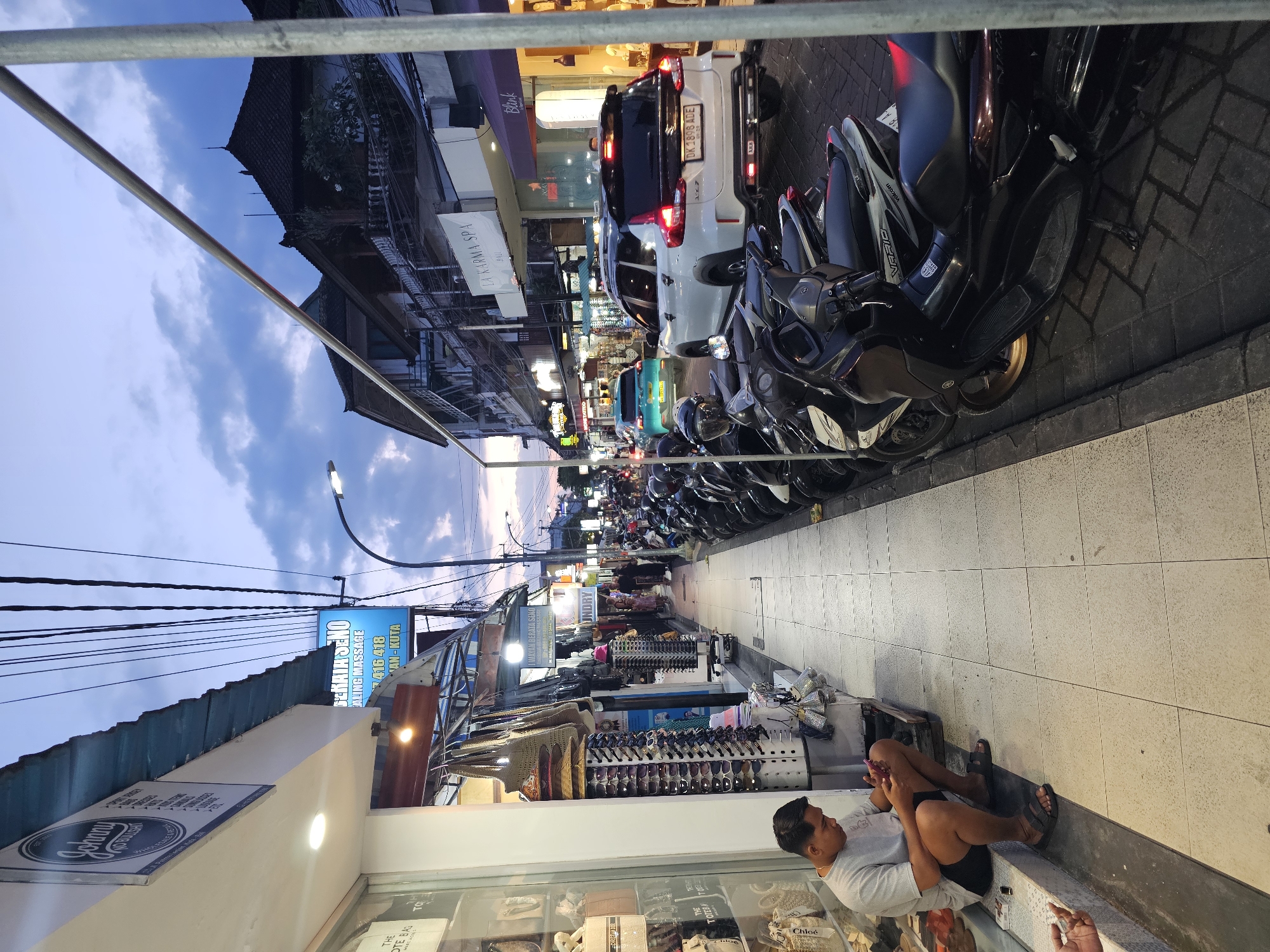 The image depicts a street scene in Kuta, Indonesia. A man is sitting on the edge of a storefront, looking at a mobile phone. The storefront displays items for sale behind glass. A blue sign reads "SEKALA SENO ALING MASSAGE" followed by "416 418" and "AN-KUTA" and "BERATA SENO". The street is crowded with parked scooters and cars. Shops and buildings line the street, and the sky is visible with a sunset.