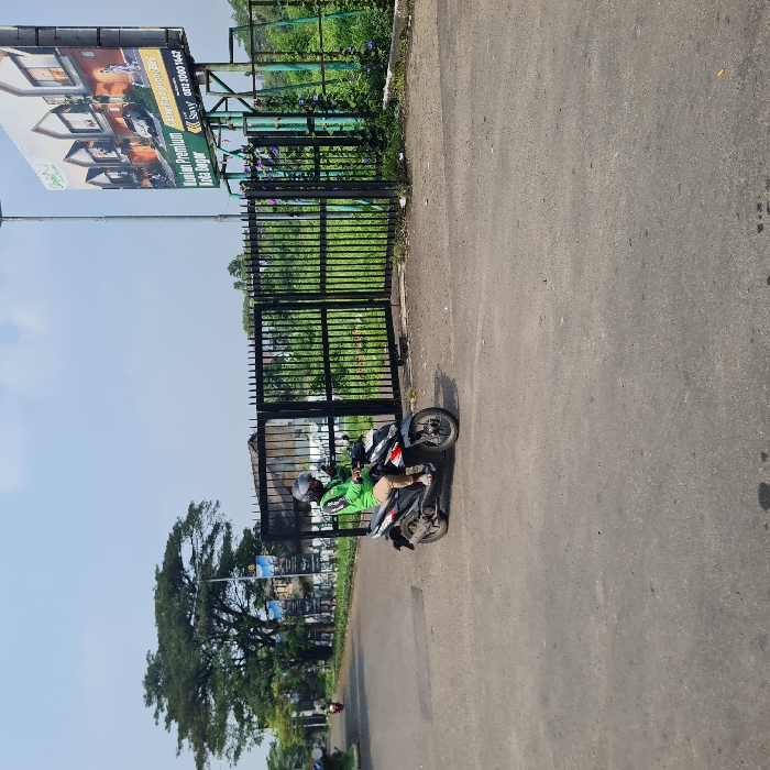 The scene is a street in Dramaga, Indonesia, with a man riding a black scooter towards the left. A black metal gate is open to the right. The scooter driver is wearing a green shirt, helmet, and gloves. The gate is closed. A large billboard advertising a residential development is on the right side of the road. The road is paved and there is grass and trees on either side of it. The sky is a light blue.