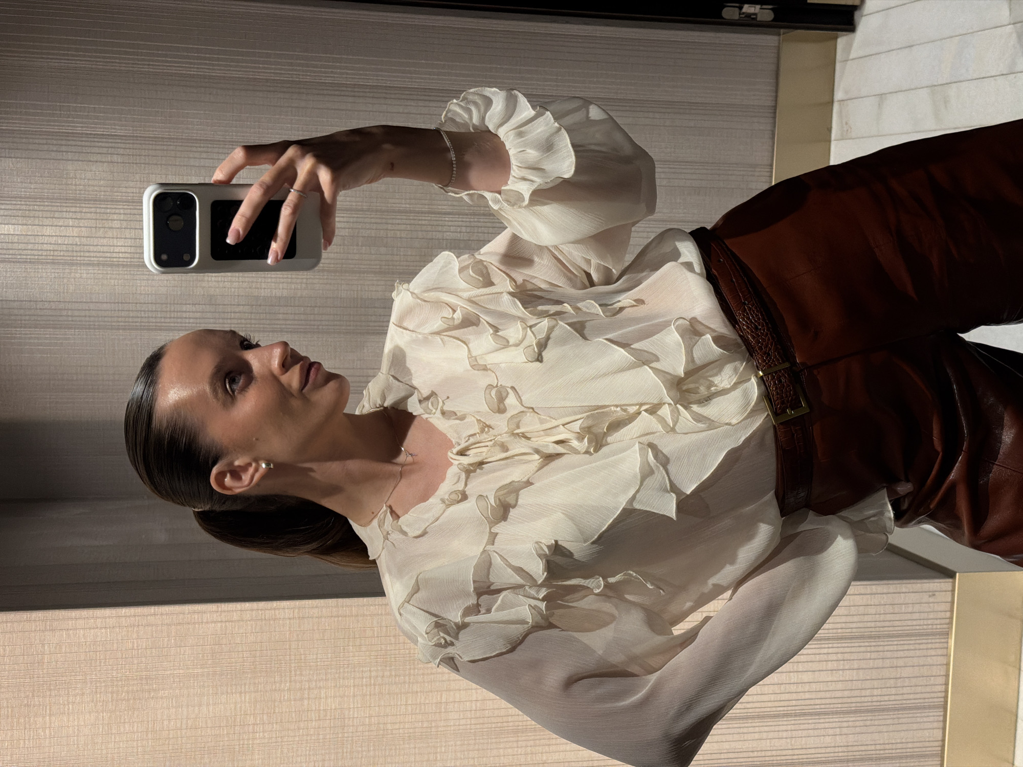 A woman with her hair in a high ponytail takes a mirror selfie. She is wearing a cream-colored ruffled blouse and brown leather pants. She is also wearing a thin necklace, earrings, and a bracelet. The background appears to be a hotel hallway with textured wallpaper and a tiled floor. The lighting is dim, casting shadows on the walls. This scene is not located in Budva, Montenegro.