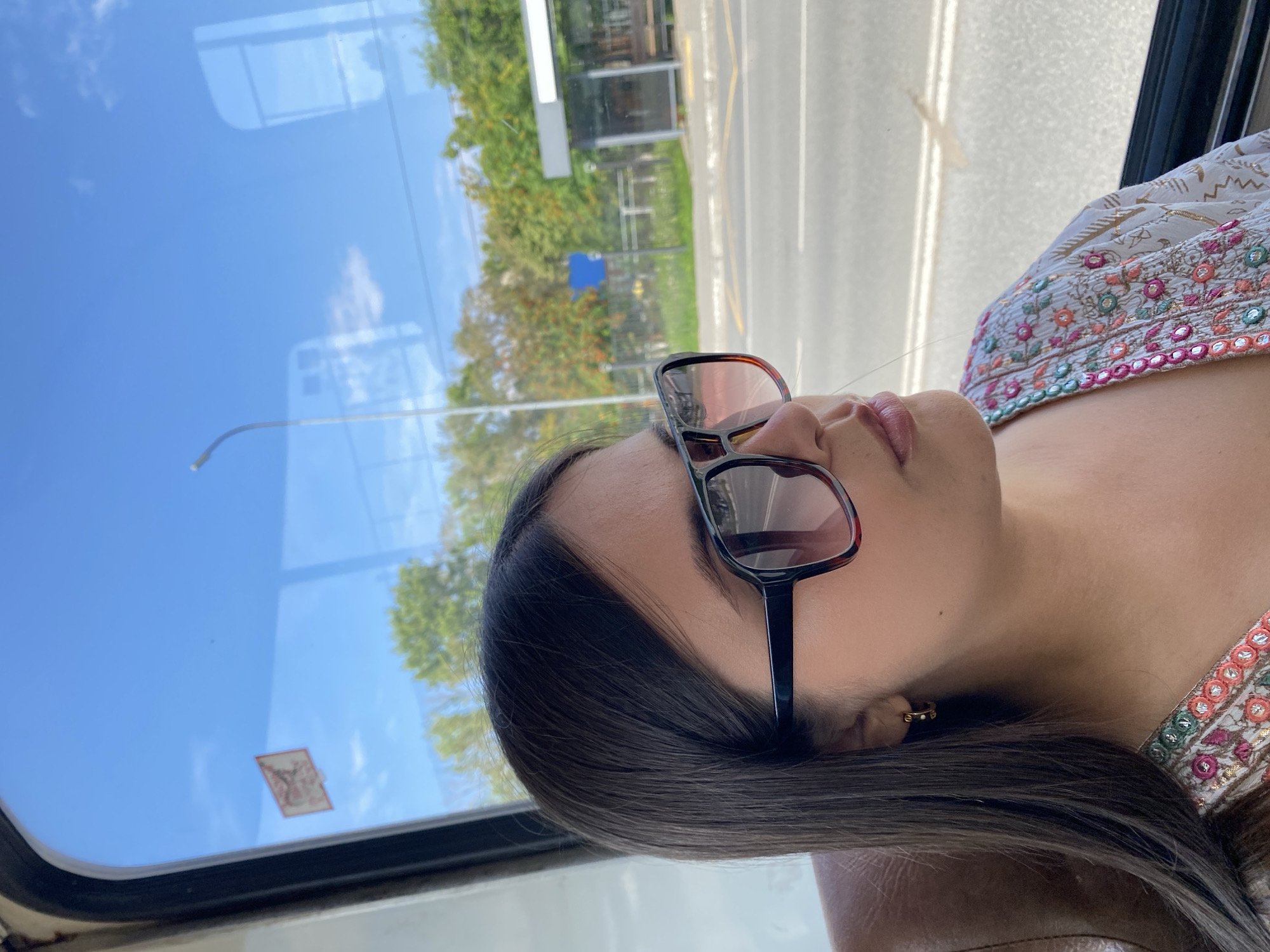 A young woman with dark hair and sunglasses looks out the window of a vehicle, likely a bus, as it travels along a street in Chaikovsky, Russia. The sun is shining, creating reflections on the glass. Outside the window, trees and a bus stop shelter are visible. The woman is wearing a white embroidered top with colorful patterns.