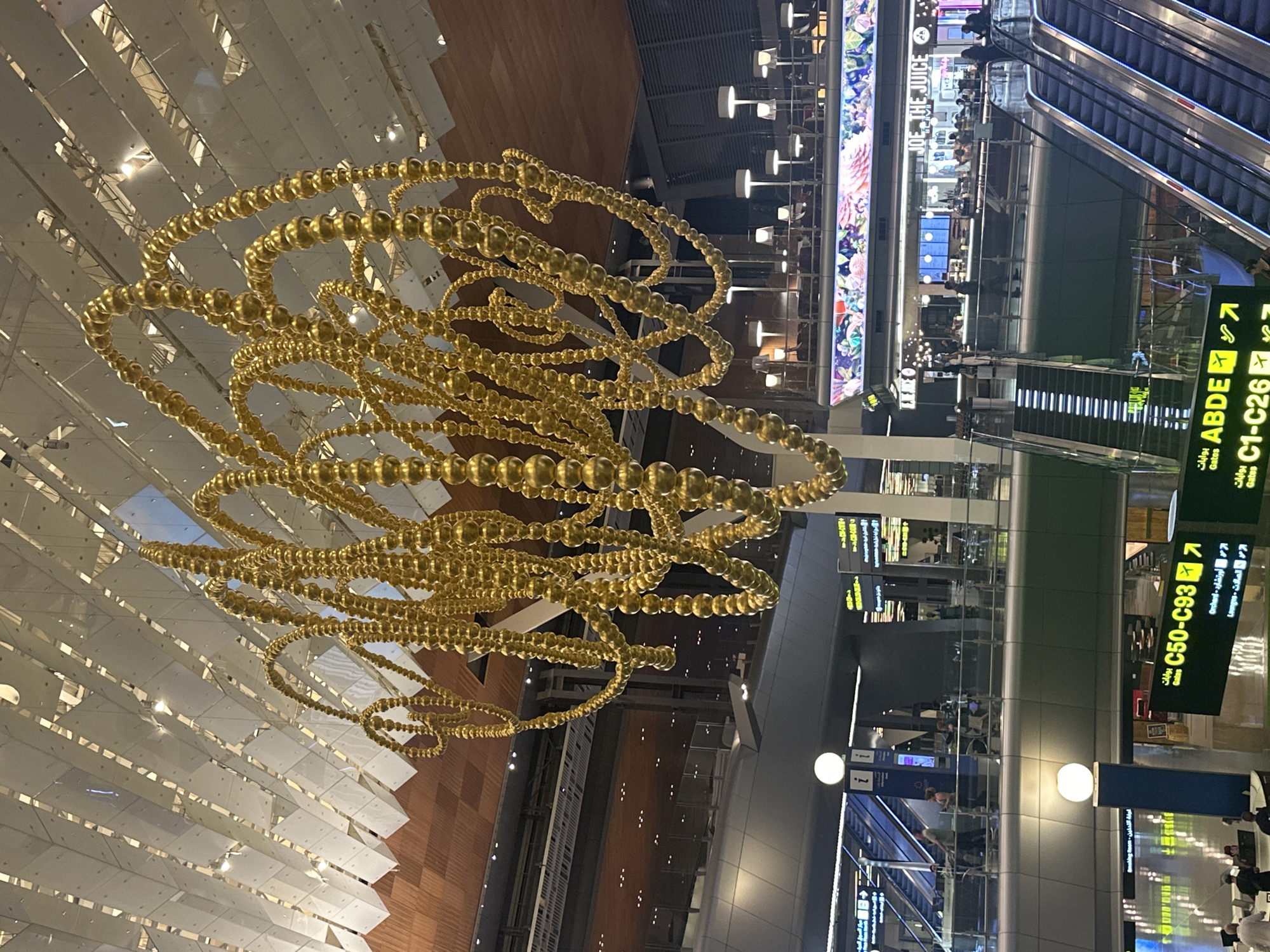 The image captures a view inside Hamad International Airport in Doha, Qatar. A large, intricate golden sculpture made of interconnected spherical elements hangs from the ceiling, dominating the upper portion of the frame. The airport's interior features a modern architectural design with a distinctive white, angled ceiling structure and exposed wooden panels. Below the sculpture, several levels of the airport are visible. People are seen walking on escalators and walkways, with shops and restaurants lining the background. Signage indicating gate numbers and destinations, such as "C50-C93" and "ABDE," are present. The overall environment suggests a busy, contemporary travel hub.