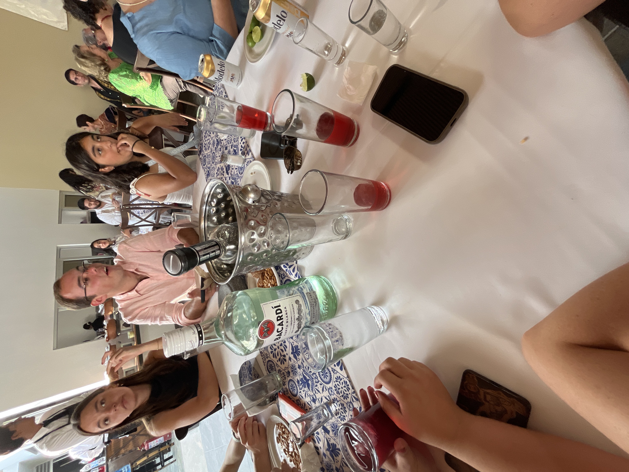 The image depicts a gathering of people around a table laden with drinks, food, and other items. There are multiple individuals visible, engaged in conversation and socializing. The table is covered with a white tablecloth and features various drinking glasses, bottles of alcohol (including Bacardi and Modelo), snacks, and what appears to be a bucket of ice. A smartphone is also present on the table.
The location is possibly San Bernardino Tlaxcalancingo, Mexico.