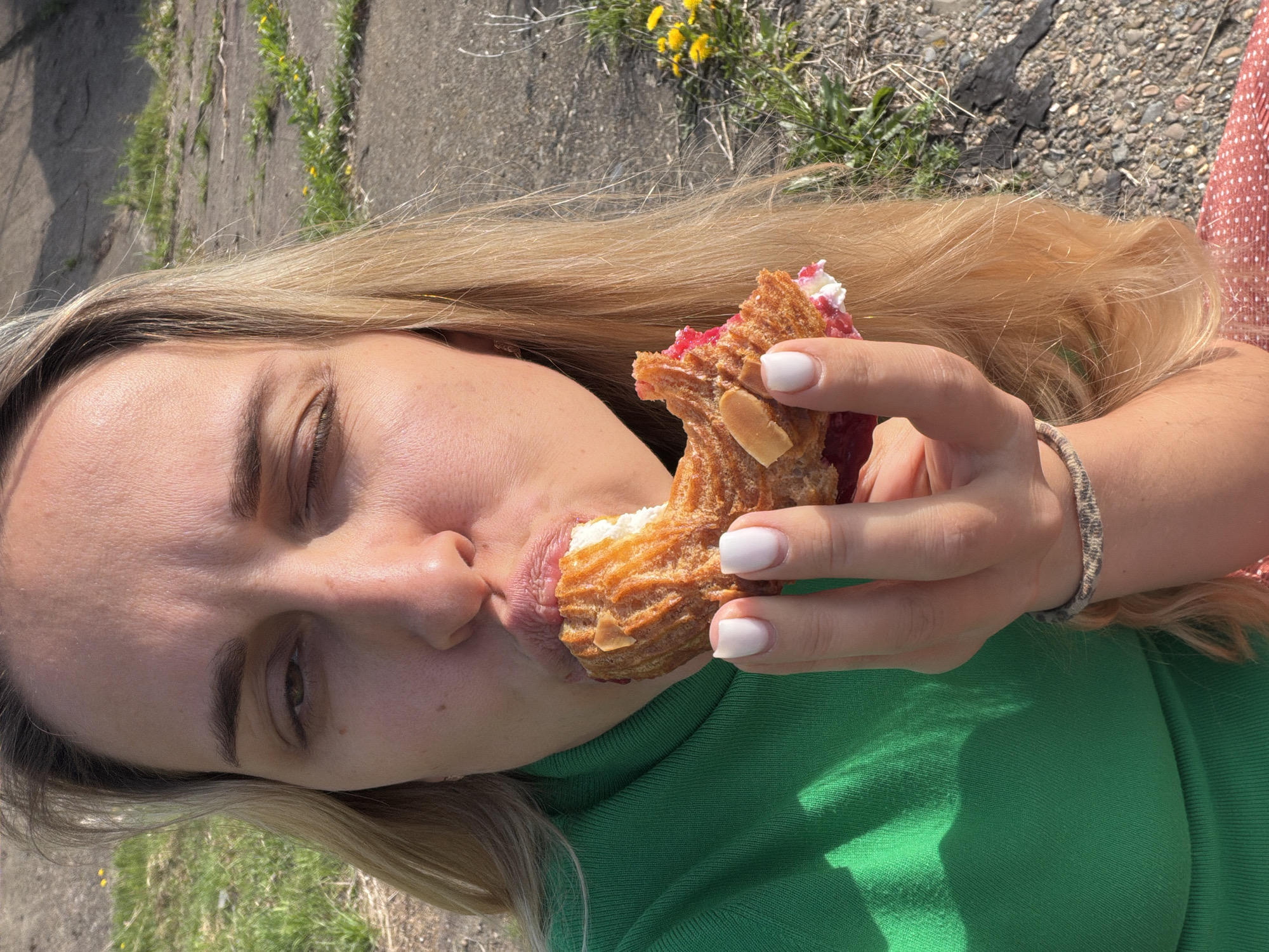 A light-skinned woman with blonde hair is eating a pastry outdoors. She is wearing a green shirt and a red and white polka dot dress. Her nails are painted white, and she is wearing a bracelet. The pastry has almonds on top and is filled with white cream and red jam. The background shows pavement, grass, and small yellow flowers. This photograph was taken in Chaikovsky, Russia.