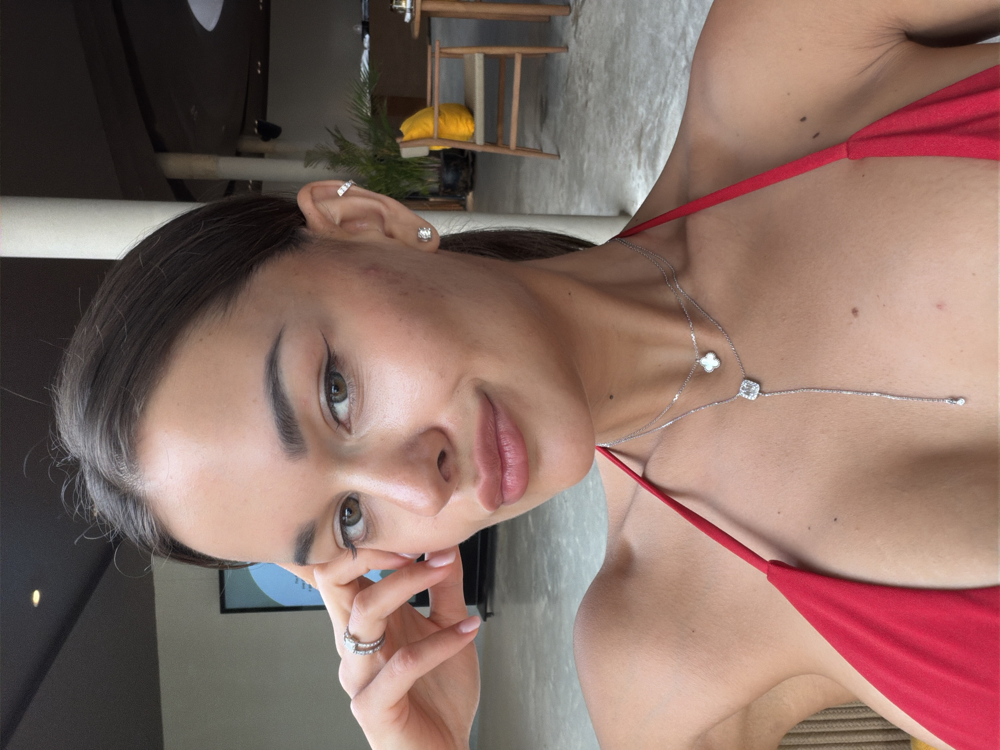 A young woman is positioned in a close-up shot. Her head is tilted to the right. She has dark hair, fair skin, and is wearing a red top. She has a silver necklace with a floral design. She also wears rings on her fingers and jewelry in her ears. The background shows a partially visible interior and some outdoor scenery, possibly a patio area. The location is potentially within Choeng Thale, Thailand, as requested. Details are limited to what is visible.