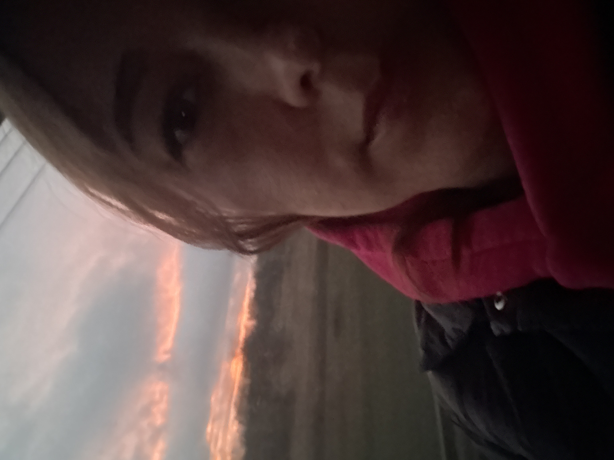 The image shows a selfie of a person with blonde hair and a red scarf or hoodie. They are partially visible, with the focus on their face. The background includes a dusky or dawn sky with orange streaks, suggesting a sunrise or sunset. Below the sky, there is a blurry landscape, likely a field or open space. The location is indicated as посёлок опытного хозяйства «Ермолино», Russia, but there are no visible markers in the image that confirm this location. There are no interactions or actions depicted, it is a static image.