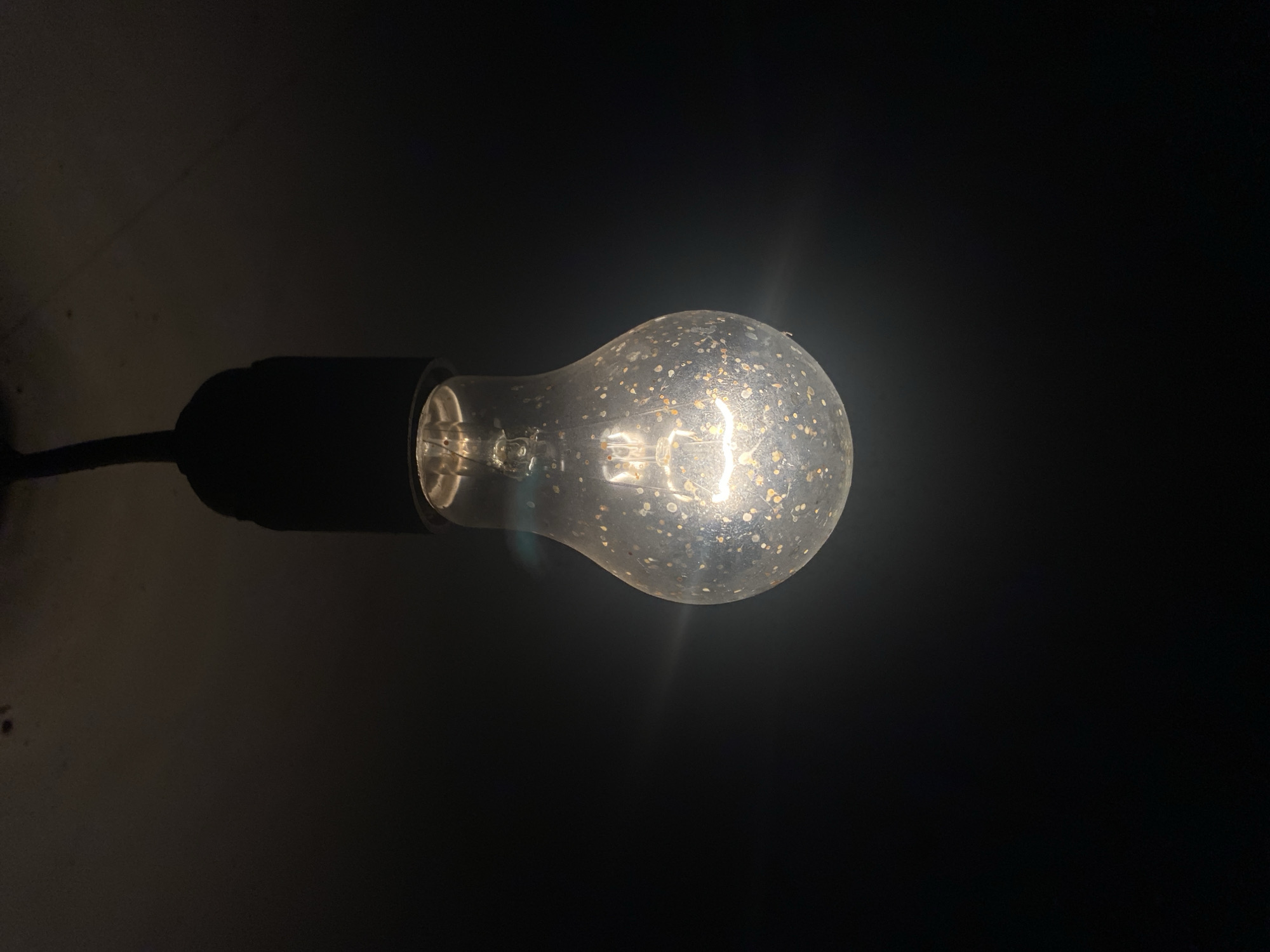 A close-up, low-angle shot features a single, incandescent light bulb suspended from a black cord. The bulb is illuminated, casting a warm glow against a dark, indeterminate background. The glass of the bulb is slightly dusty or speckled with small, irregular marks, giving it a textured appearance. The filament within the bulb is visible and glowing brightly. The top of the bulb connects to a black socket, which is attached to the cord. The overall scene is dimly lit, with the light bulb being the primary source of illumination. There is no evidence of any specific location in Kisliakovskaya, Russia, or any people present in this image.