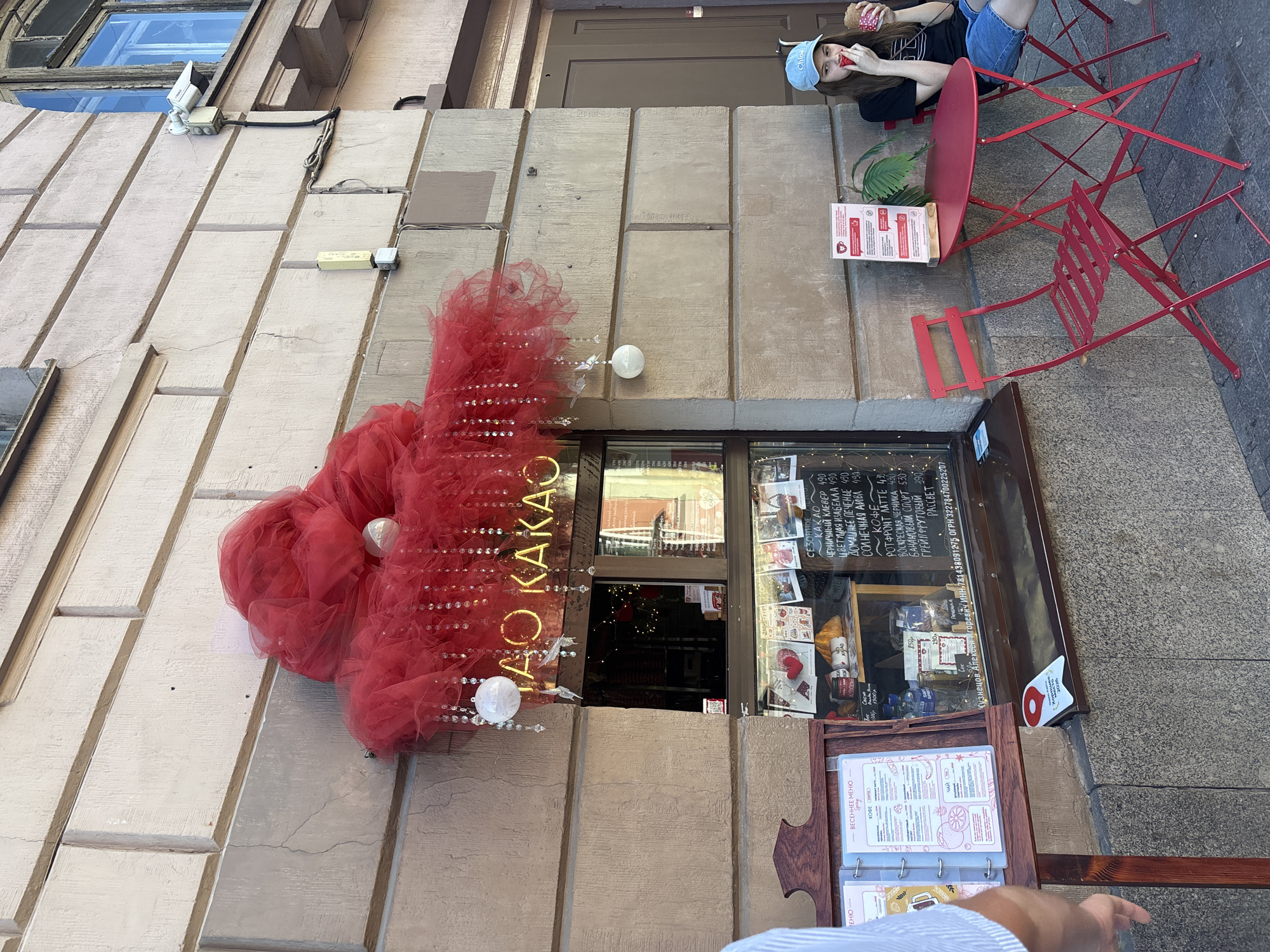 The scene depicts a daylight street view of a small cafe storefront in Saint Petersburg, Russia. The building facade is constructed of light-colored, roughly rectangular blocks, exhibiting minor cracking.
Above the central glass display window, large red tulle or netting decorations are affixed, resembling a voluminous cloud. From these decorations, multiple strings of clear beads and several white spherical ornaments, along with small white bird figures, hang downwards. Neon or illuminated Cyrillic letters spelling "САО КАКАО" (CAO KAKAO) are partially visible behind and below the red decorations.
The shop window displays various handwritten menus and price lists in Cyrillic, detailing items such as "КАКАО" (Cacao), "КОФЕ" (Coffee), and "ЧАЙ" (Tea), with prices ranging from 430 to 530. Below the window, a white sign with a red "9" and "Хорошие Кафе 2022" (Good Cafes 2022) is displayed. Further details on the window list "ИНН" (INN) and "ОГРН" (OGRN) numbers and the name "КУЗНЕЦОВ АЛЕКСЕЙ ИГОРЕВИЧ", indicating Russian business registration.
To the left of the window, a wooden stand holds an open menu book, displaying "ВЕСЕННЕЕ МЕНЮ" (Spring Menu) for coffee and tea. A partially visible individual, likely male, wearing a light-colored shirt, has their left arm extended towards this menu stand.
To the right of the window, a brown wooden door is set into the facade. In front of this door, a red oval metal folding table is placed, accompanied by two red metal folding chairs. Seated at the table on the right chair is a young female, appearing to be in her late teens or early twenties. She wears a dark t-shirt, blue denim shorts, and a light blue baseball cap with "Chloe" embroidered in white. She is holding a reddish-brown cup or cone and bringing a small item to her mouth with her right hand. A small vertical menu stand with red text on a white background, featuring a coffee cup icon, is positioned on the table, partially obscuring a potted plant.
Above the right side of the storefront, a white security camera is mounted on the building facade, pointing downwards. Below it, a yellow rectangular electrical box and exposed wiring are visible. The ground is paved with dark, possibly asphalt or stone, material. The bright, direct lighting suggests it is daytime.