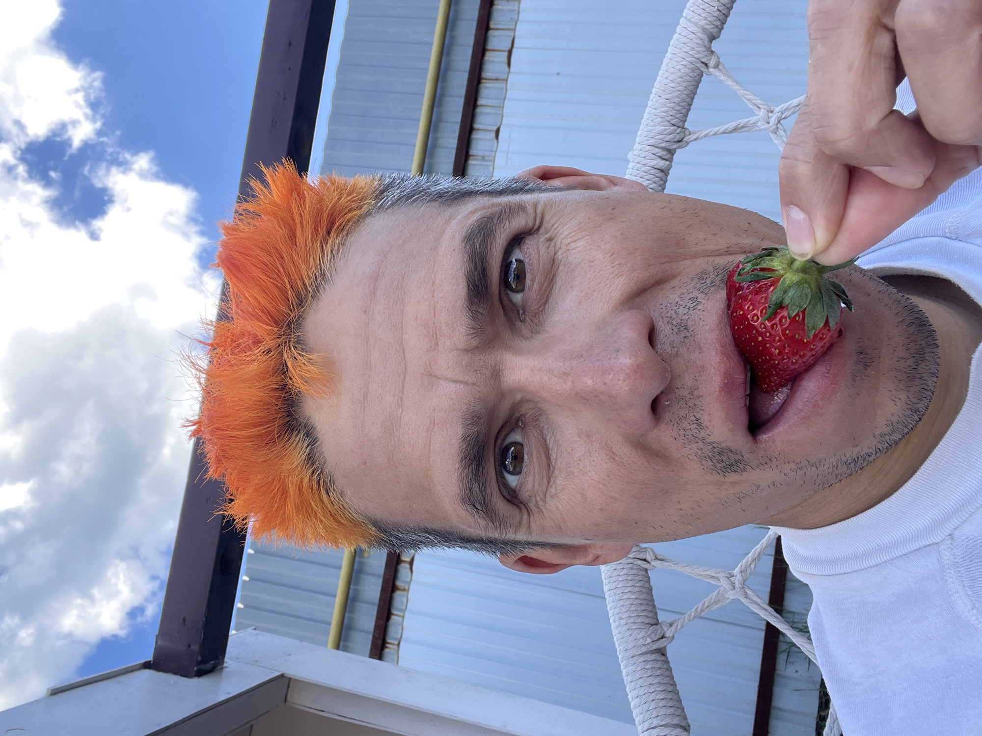 A person with orange hair is holding a strawberry in their mouth. The person is wearing a white shirt. The background appears to be the exterior of a building. There are light blue panels and a rope structure partially visible. The sky is visible in the upper left corner. There is no verifiable information about the city of Chaikovsky, Russia.