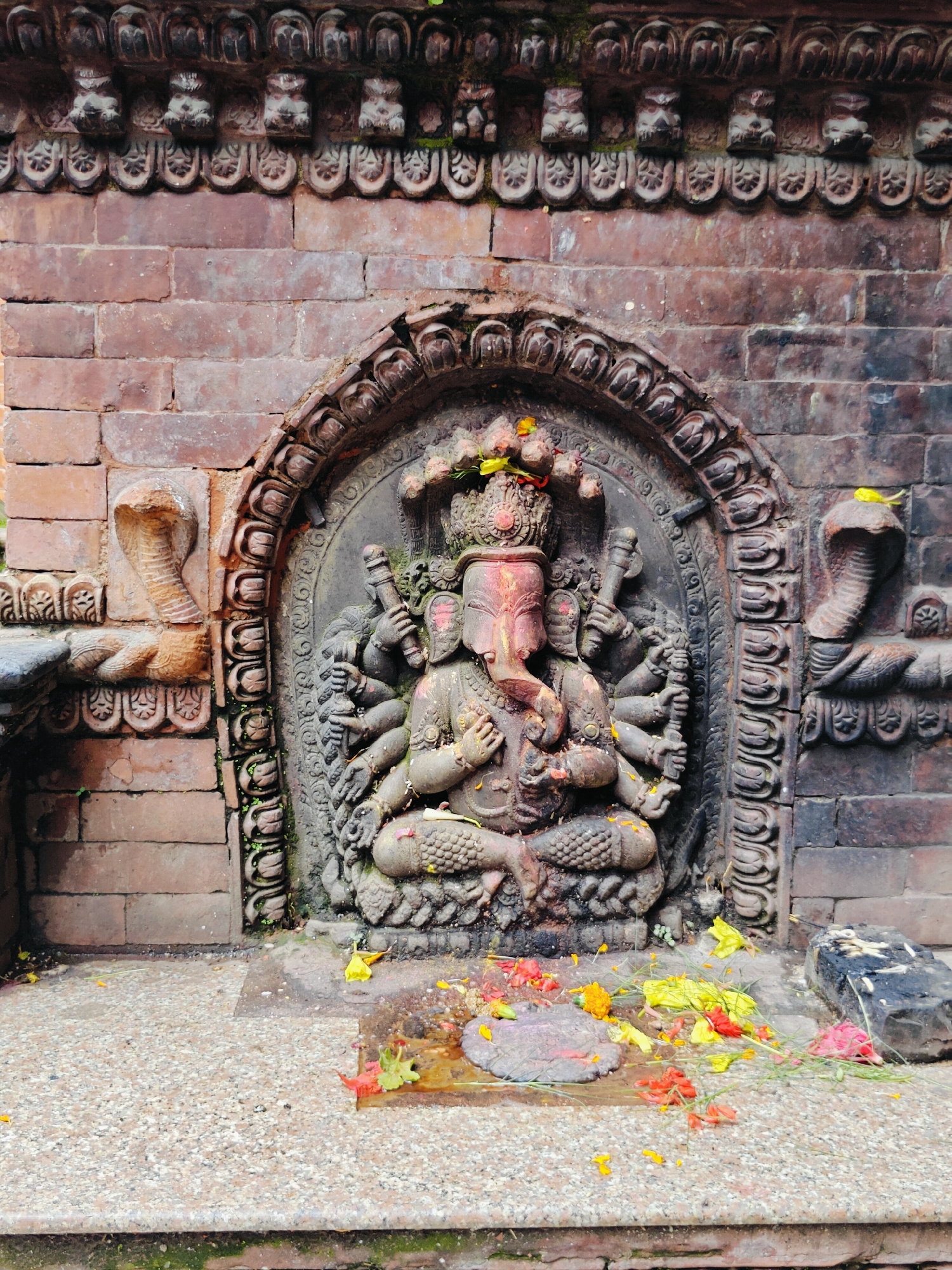 An outdoor, low-angle, medium shot captures a stone sculpture of Ganesha, the Hindu deity with an elephant head, seated in front of a brick wall in Lalitpur, Nepal. Ganesha is depicted with multiple arms, each holding an object, and a crown adorns his head. The sculpture is richly decorated with intricate carvings. Flanking Ganesha on either side are carved serpents. The stone base in front of the sculpture is adorned with fallen yellow, red, and pink flowers, indicating a recent offering or ritual. The surrounding brickwork is old and weathered, with moss and dirt accumulating in crevices. The ground in front of the stone base is a rough, speckled concrete surface, with a small dark stone block to the right of the image. The overall scene suggests a place of worship or a historical site.