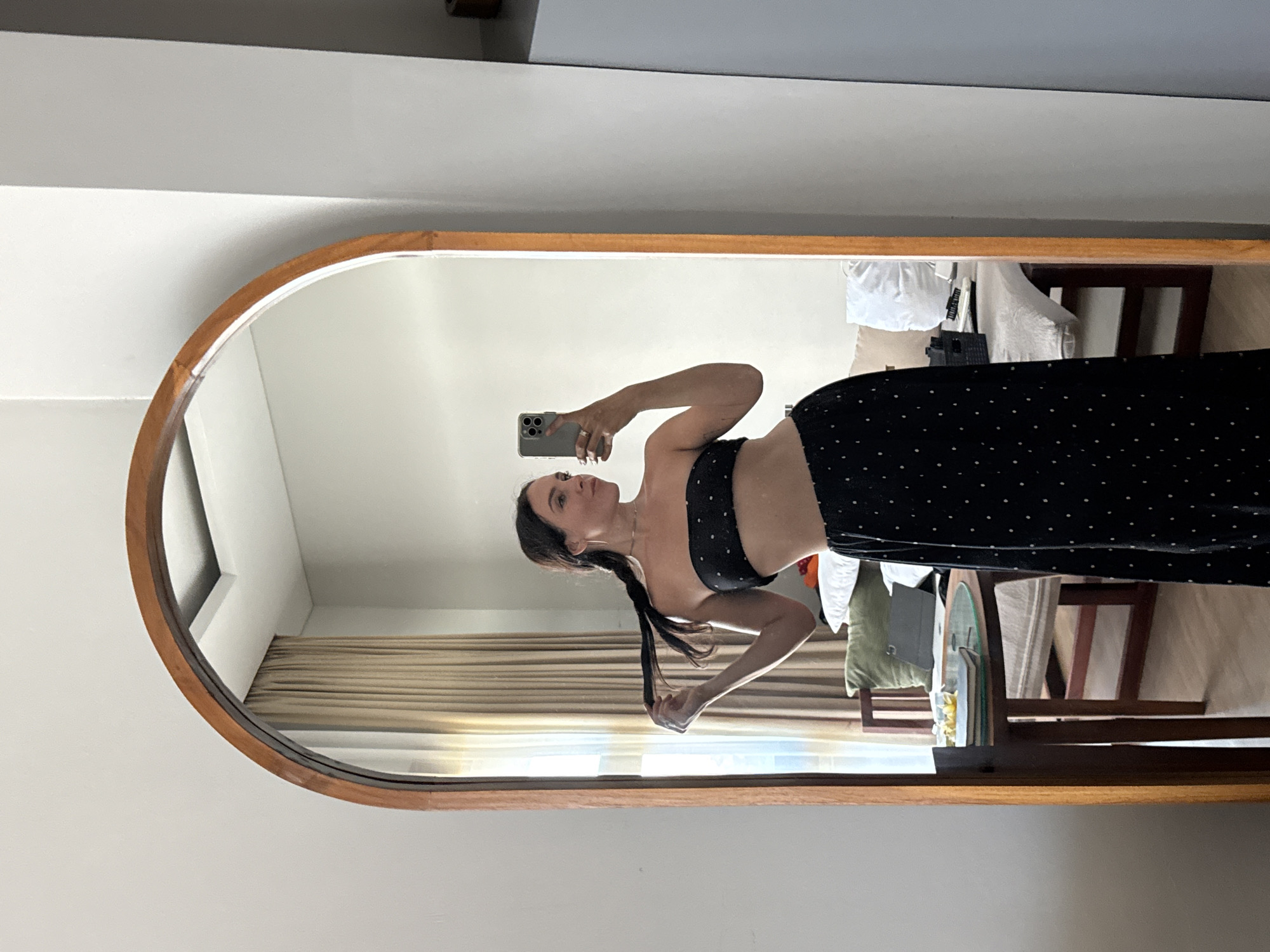 A woman stands in front of a mirror, holding her phone to take a selfie. She is wearing a black bandeau top with white polka dots and a matching high-waisted black skirt with white polka dots. Her dark hair is in a braid. The mirror has a wooden frame and is tall and arched at the top. The reflection shows a room with a window covered by beige curtains, a small round table with a book and some yellow items, and a chair. The environment appears to be an indoor residential setting, possibly a hotel room in North Kuta, Indonesia, given the decor and lighting.