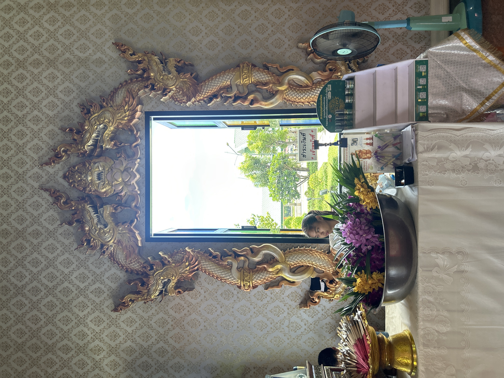 In Thai Ko Subdistrict, Thailand, a young woman with her hair pulled back is visible through an ornate, gold-trimmed doorway featuring dragon motifs. She is looking to her right. The doorway opens to an exterior scene of lush greenery and trees under a bright sky. Inside, the wall is decorated with a repeating pattern. In the foreground, a table is draped with a white lace-trimmed cloth. On the table sits a large silver bowl filled with purple and yellow flowers, and to its left, a golden stand holds numerous incense sticks. To the right of the doorway, a white tiered display stand holds several small cylindrical items, and a blue oscillating fan is positioned nearby. A sign with Thai script is visible on the stand.