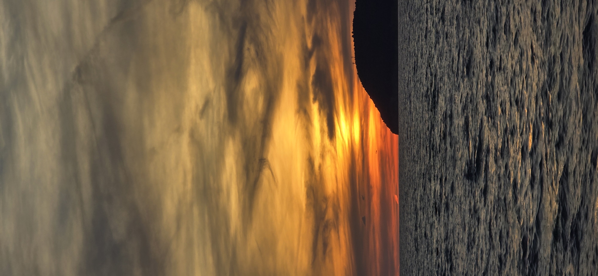 The image shows a sunset in Bar, Montenegro. To the left of the image are wispy gray and orange clouds. To the right of the clouds is a dark, silhouetted mountain. To the right of the mountain is the dark gray surface of the Adriatic Sea with visible reflections of the sun.