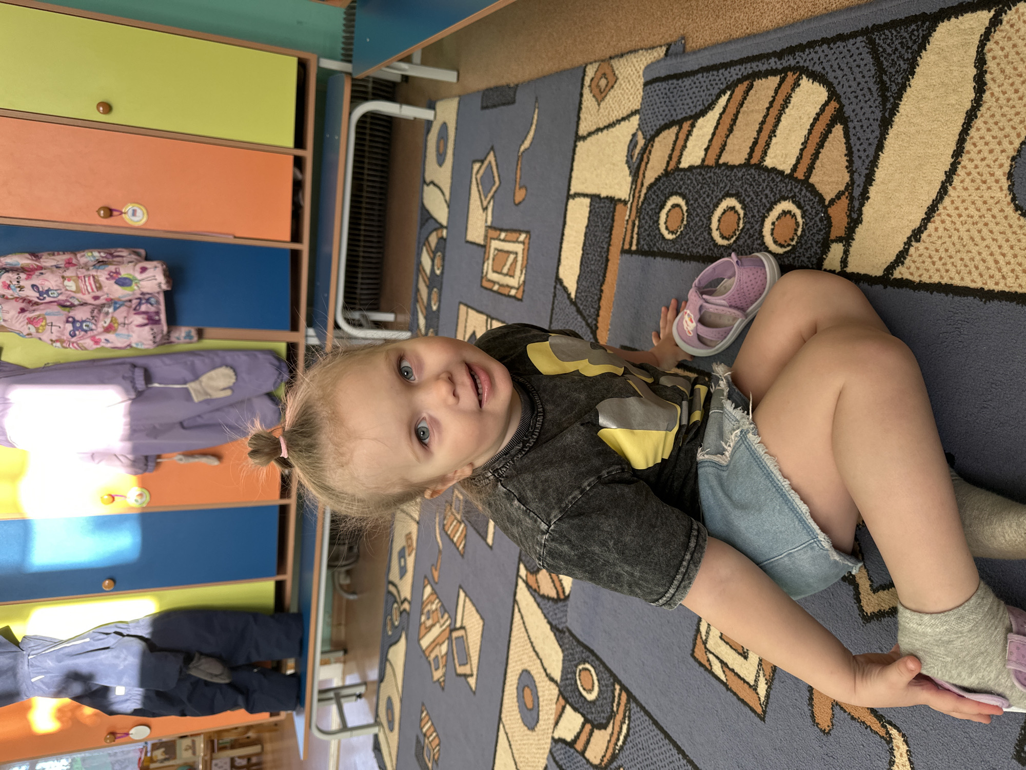 A young child with blond hair pulled into a small ponytail sits on a colorful rug in what appears to be a playroom in Прикамский, Russia. The rug has a pattern of cartoon-like vehicles. The child is wearing a gray shirt with a graphic, denim shorts, and gray socks. Nearby is a pair of purple sandals. Behind the child is a set of lockers or cubbies with doors in various colors: yellow, orange, and blue. Hanging on the lockers are various jackets and clothing items. In the background, there is what appears to be a metal frame, possibly part of furniture or a play structure.