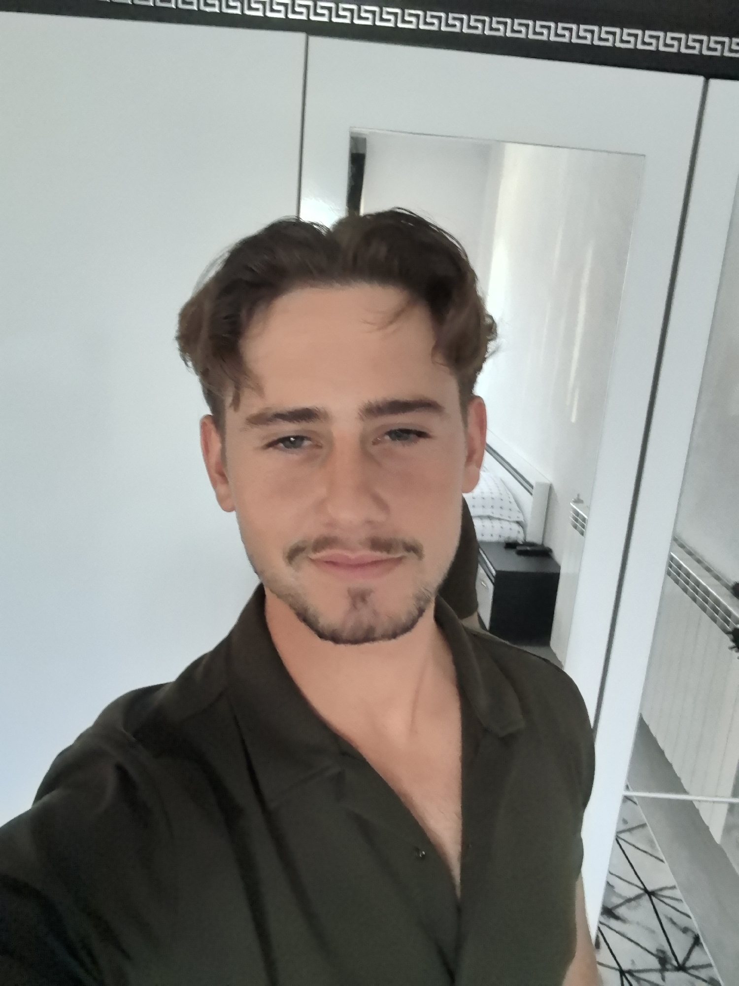 A man is standing indoors, seemingly taking a selfie. He has brown hair styled with a slight wave, light eyes, and a neatly trimmed beard and mustache. He is wearing a dark green collared shirt with a button closure. The background includes a white wardrobe or closet with a decorative geometric border at the top. A mirror is partially visible, reflecting a room with a bed, a bedside table, and what appears to be a radiator. The location cannot be determined as Skrebatno, Bulgaria from the image.