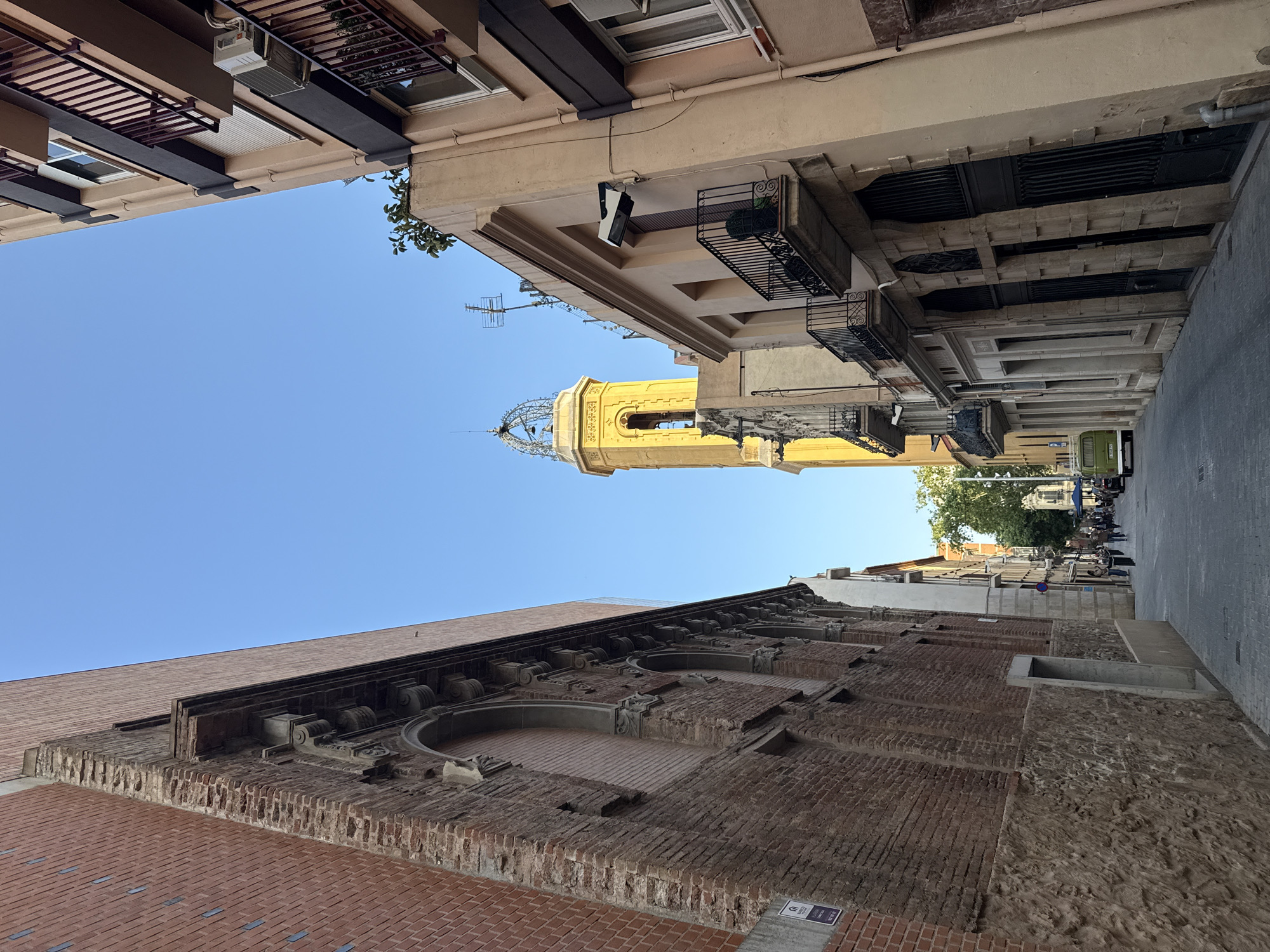 A ground-level shot looking up at a narrow city street in Barcelona, Spain, with tall buildings on either side. The buildings are a mix of architectural styles and materials, including brick, stone, and stucco. The buildings are tightly packed together, with balconies and other architectural details visible. A yellow church tower is centered in the frame between the buildings. The sky is blue and clear. Some people and vehicles are visible in the distance.