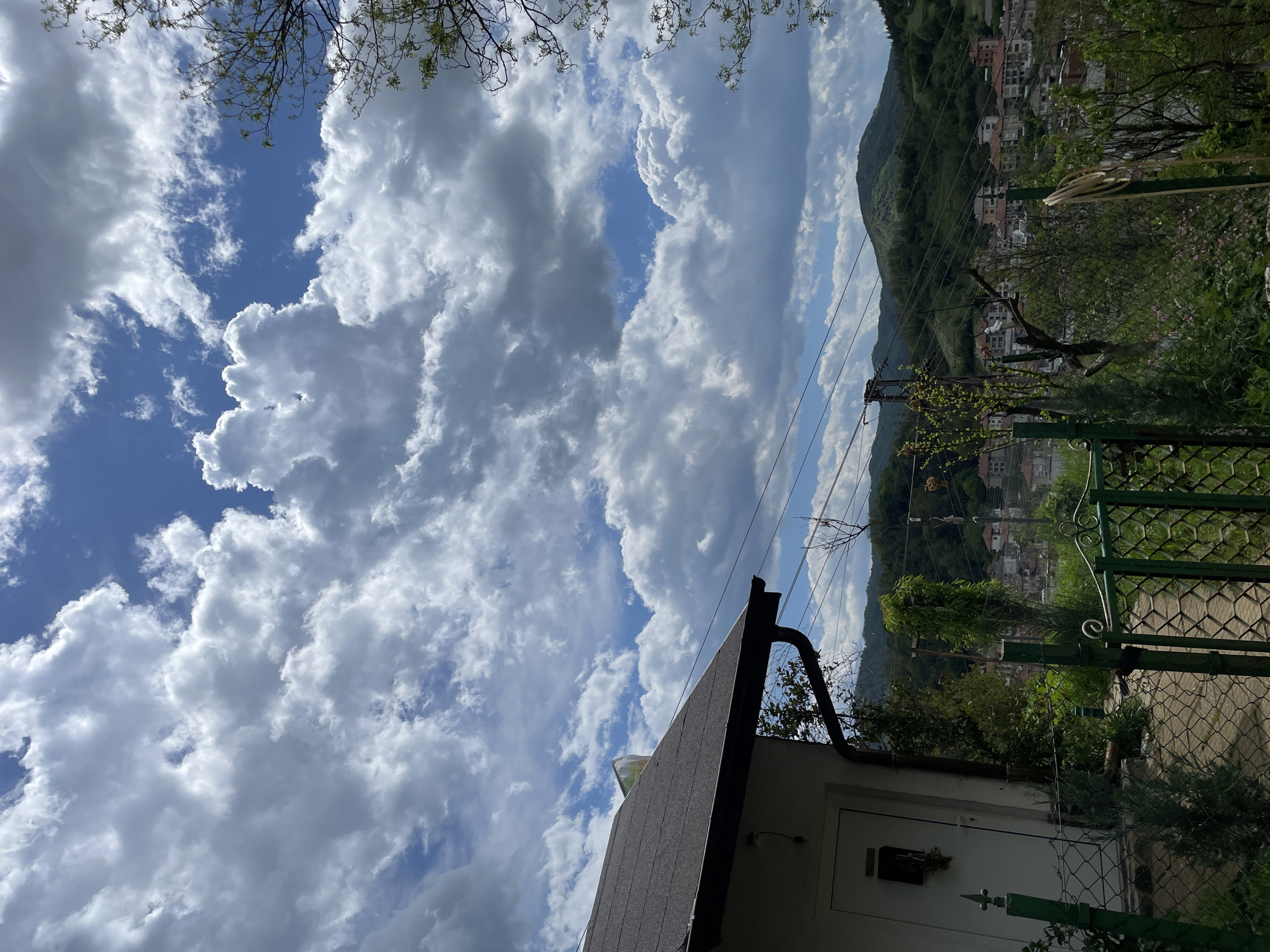 A view from Zlatograd, Bulgaria, shows a bright, partly cloudy sky with cumulus clouds filling the upper portion of the frame. Below, a sloped roofline of a building with dark shingles and a black gutter runs diagonally across the lower left of the frame. The white wall of the building is partially visible beneath the roof. To the right, a green metal fence is partially visible, with lush green vegetation and trees extending beyond it. In the distance, a hillside covered in green foliage slopes down to a town with buildings clustered together. Power lines stretch across the scene, intersecting with the sky and mountain. The view is angled upwards, giving a sense of looking up from a lower vantage point.