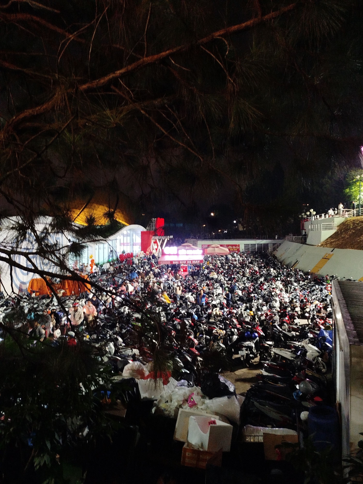 The image depicts a large outdoor gathering at night in Da Lat, Vietnam, likely a festival or event, with an enormous crowd of parked motorcycles filling a significant portion of the scene. The motorcycles are densely packed together, forming a sea of vehicles. In the background, a white tent-like structure with illuminated signage is visible, suggesting a temporary venue or stall. Numerous people are present, milling about amongst the motorcycles and near the illuminated structure. The environment appears to be a public space, possibly a street or a designated event area, with some foliage in the foreground obscuring parts of the view. The lighting is predominantly artificial, with bright lights from the event structures and the glow of various motorcycle headlights. The overall impression is one of a vibrant and crowded nighttime event.