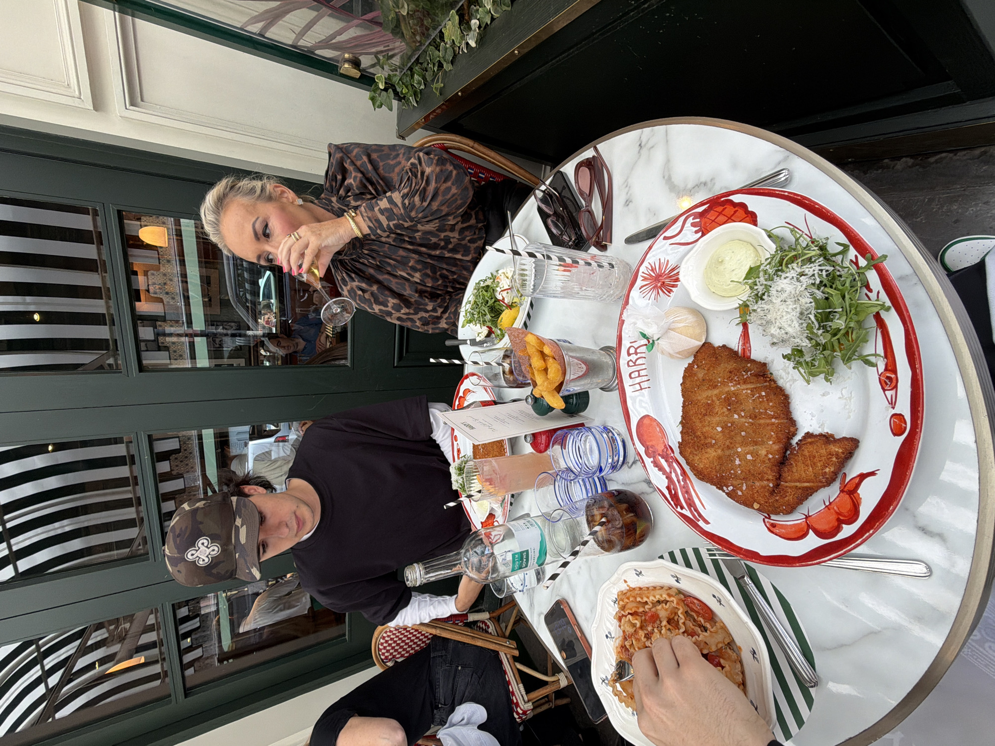 The image shows a table at a restaurant with two people seated. The person on the left, a male, is wearing a camouflage baseball cap and a black t-shirt. The person on the right, a female, is wearing a leopard-print top and jewelry, and is drinking from a glass. On the table are plates of food, including what appears to be pasta and a breaded cutlet with salad and a dipping sauce, as well as drinks and cutlery. In the background, there are windows with black and white striped awnings. The setting appears to be an outdoor dining area. The restaurant's name, "HARRY'S", is visible on one of the plates. This detail, along with the striped awnings and the overall aesthetic, suggests the location is Harry's Bar in the city of London, United Kingdom.