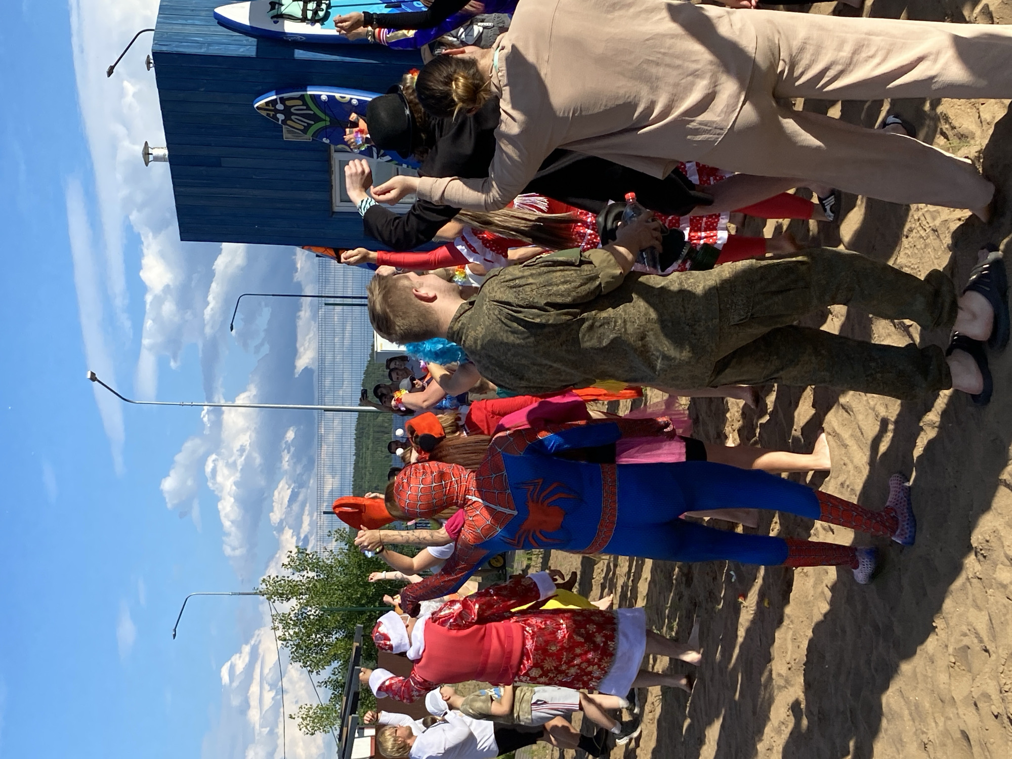 The image shows a group of people on a sandy area with a blue building in the background. The people are dressed in various outfits, including what appears to be a Santa Claus costume, a Spider-Man costume, and other casual wear. Some individuals are holding objects, and many have their arms raised. In the background, there are surfboards leaning against the blue building. The environment appears to be outdoors, with a clear blue sky and some clouds. The presence of surfboards and the sandy terrain suggest a location near water, potentially a beach or a recreational area. The overall scene depicts a gathering or an event, possibly a festival or a party. The location is identified as the city of Chaikovsky, Russia.