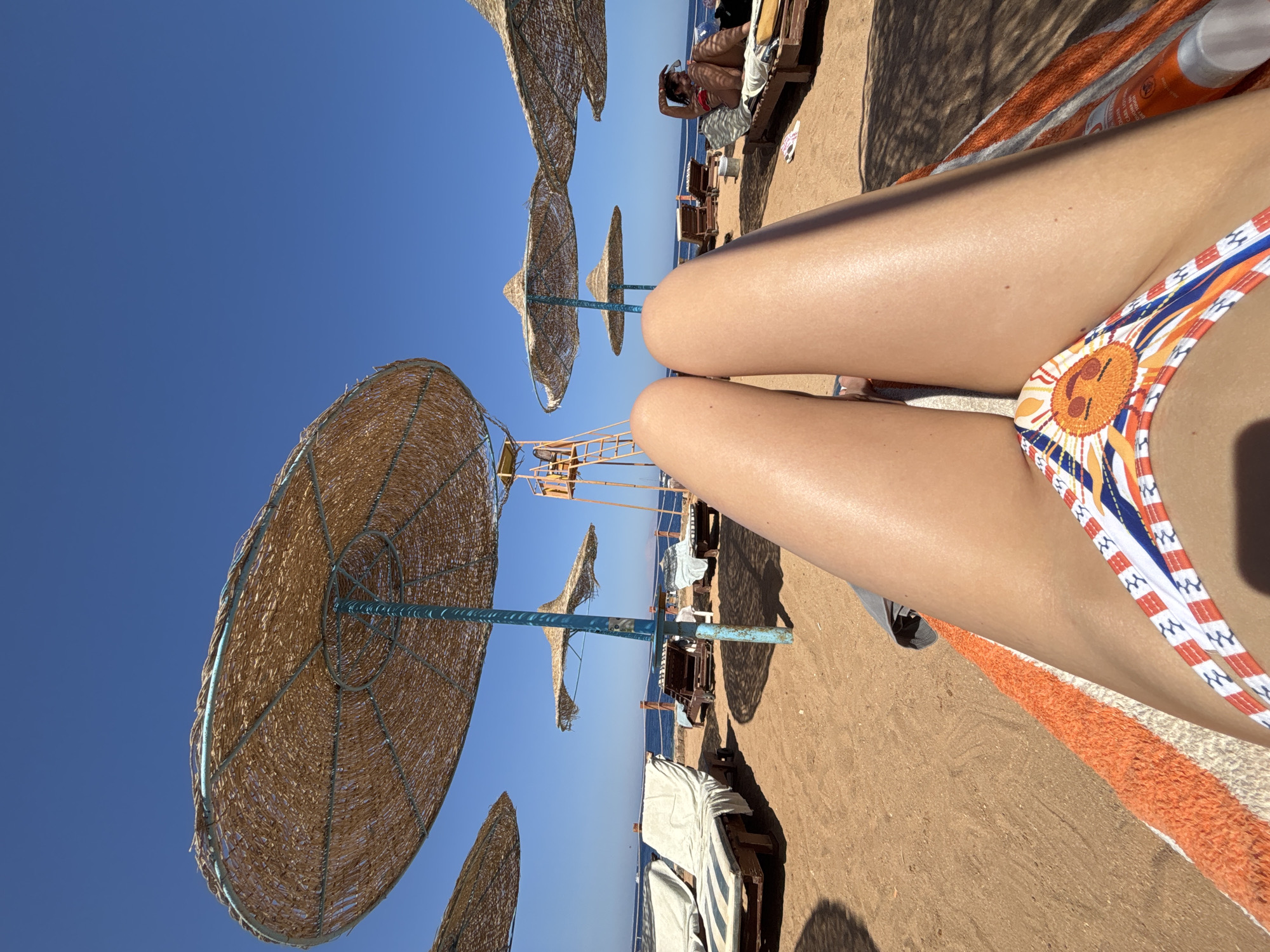 The image shows a beach scene. There are multiple straw umbrellas shading lounge chairs on the sand. The sky is clear and blue. In the foreground are the legs of a person in a patterned bikini. The location is a beach.