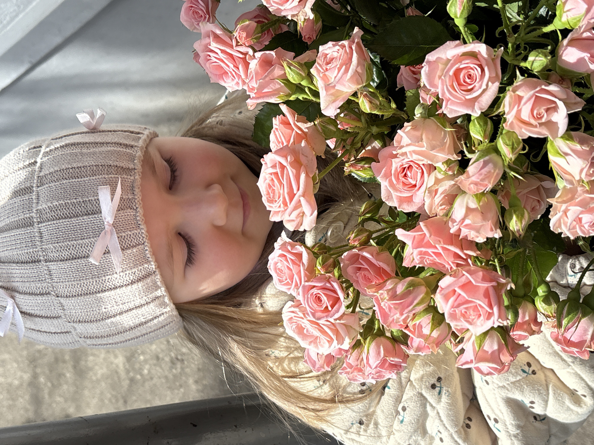 The image, oriented vertically, captures a young child lying down with a large bouquet of light pink spray roses. The child's head is positioned towards the lower left of the frame, with the mass of flowers filling the right side. The child, likely a girl, has fair skin and light-colored hair, and her eyes are gently closed, conveying a peaceful and serene expression. She wears a light beige knitted hat decorated with small pink and white ribbon bows, and part of a light-colored, subtly patterned garment or blanket is visible beneath the flowers. The abundant bouquet consists of numerous light pink roses in various stages of bloom, from tight buds to fully open flowers, interspersed with dark green leaves. The scene is bathed in bright natural sunlight, creating strong highlights and soft shadows. The background behind the child's head appears to be a light-colored paved surface, like concrete, and a dark, horizontal structure, possibly a railing or bench, is visible in the lower left. The setting is outdoors in Chaikovsky, Russia, during the daytime. No text is visible in the image.