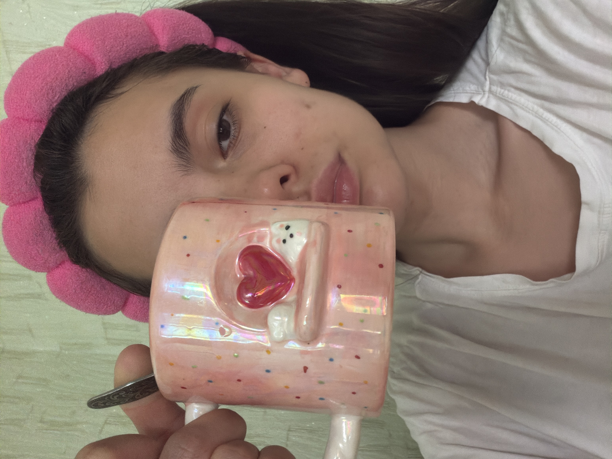 The image shows a young person holding a pink, iridescent mug with colorful speckles and a 3D design of a heart and a white cat-like figure. The person is wearing a pink, puffy headband and a white t-shirt. Their face is partially obscured by the mug. The background appears to be a wall with a textured surface. The image was taken in Chaikovsky, Russia.