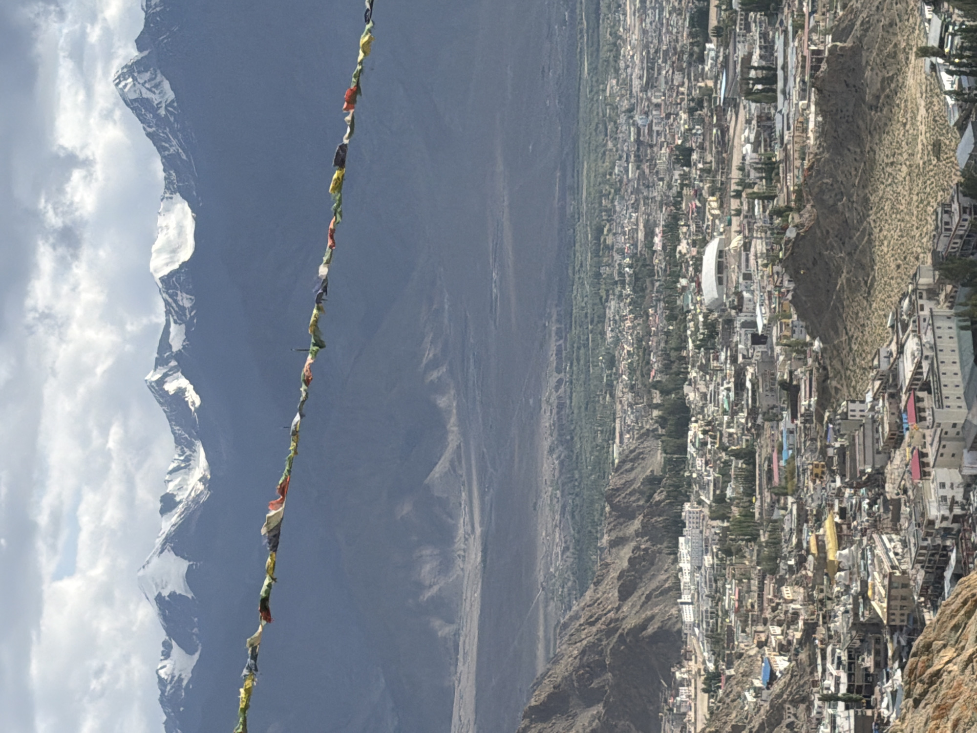 The image captures a panoramic view of Leh, India, with a mountain range in the background and prayer flags strung across the foreground. The prayer flags, colorful and weathered, are a prominent element, suggesting a spiritual or cultural context. Below the flags, the rugged terrain of the Himalayas is visible, with snow-capped peaks piercing the cloudy sky. The mid-ground features a vast, arid landscape, transitioning into a densely populated city. The city of Leh is depicted with a mix of traditional and modern architecture, characterized by flat-roofed buildings, monasteries with golden spires, and winding streets. The overall impression is one of a high-altitude settlement nestled amidst a dramatic mountain setting, conveying a sense of remoteness and cultural richness.