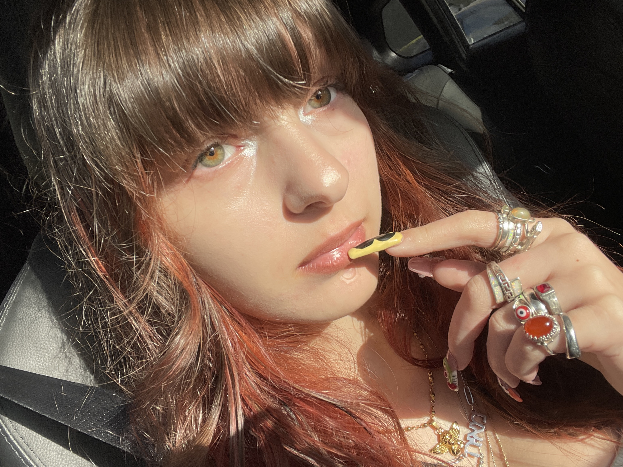 A close-up, sunlit portrait features a young woman seated inside a car. Her long, reddish-brown hair, styled with bangs, cascades around her face, catching strong highlights from bright daytime sunlight. She possesses striking green-hazel eyes and glossy lips, which she gently touches with the index finger of her right hand. This hand is adorned with numerous silver rings, including those set with carnelian, opal, and a red gem, alongside an evil eye design. Her long, manicured nails display intricate designs, notably a yellow nail with a black pattern. Around her neck, multiple delicate necklaces are visible, one featuring a "CM" pendant and another a winged charm. The background subtly shows elements of a dark car interior, including a black seat and a black seatbelt. The strong, direct sunlight suggests a clear, sunny day. The setting is a car, located in Raanana, Israel. Her gaze is direct and composed.