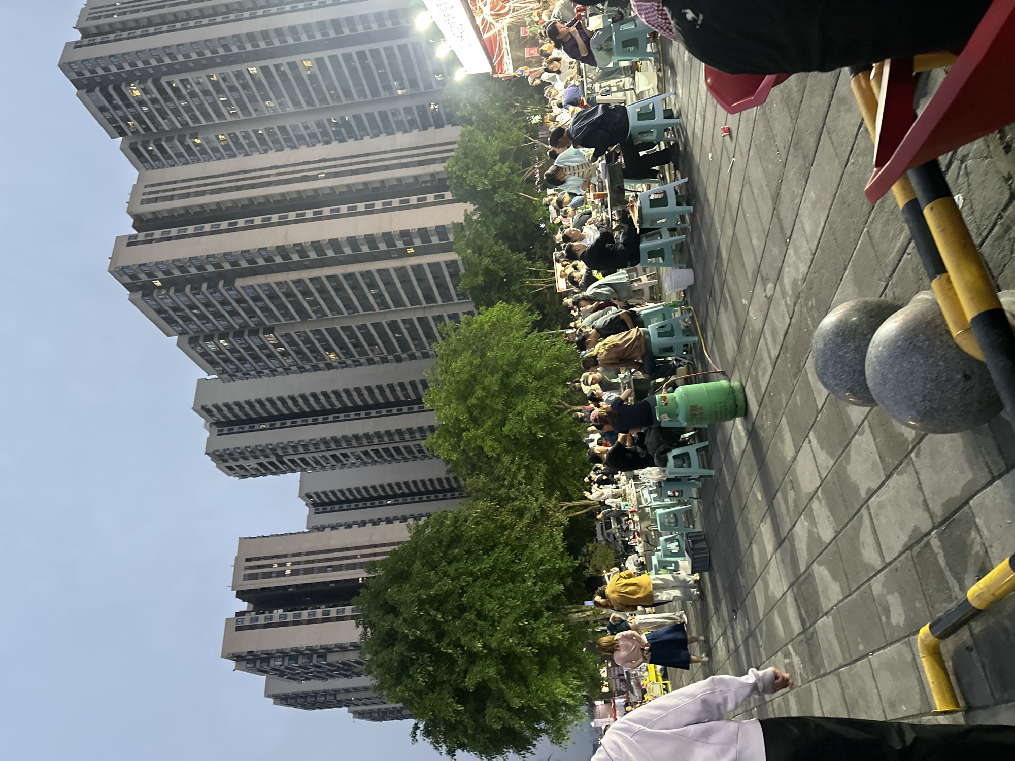 The image captures a bustling scene in Chongqing High-tech Zone, China. A tall, modern residential building with numerous stories dominates the background. Along the street, a line of people sits at small, light blue plastic tables and chairs, engaged in eating and socializing, possibly at a street food market. Lush green trees partially obscure the view of the building, adding a touch of nature to the urban landscape.
At the foreground, pavement stones cover the ground. A blurred, red and yellow barricade structure stands on the right. Part of a person's white sweater, black pants, and hand is visible in the lower foreground, suggesting their proximity to the scene. A green gas tank sits on the ground near the tables. The sky is a pale blue, indicating either early morning or late evening.