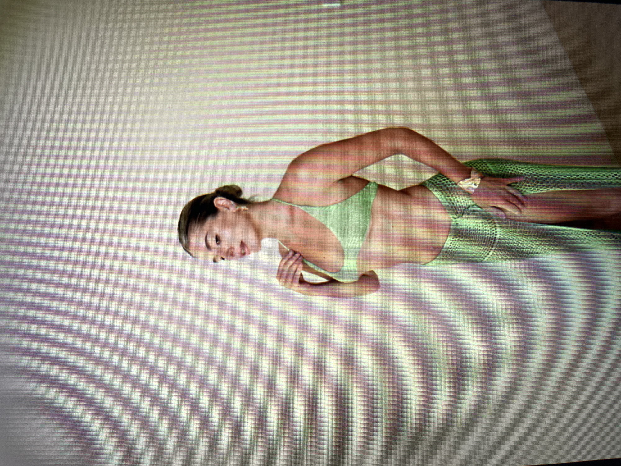 The media file shows a person, appearing to be a young woman, posing in a studio setting. She is wearing a light green, knitted bikini top and a matching knitted skirt with a slit up the side. The woman has her hair pulled back in a bun and is wearing a gold earring and a gold watch. She is turned to her left, with her left arm bent at the elbow and her hand resting on her chest. Her head is turned to her left, and she appears to be looking slightly downwards. The background is a plain, neutral-colored wall, suggesting an indoor photoshoot. The lighting is soft and even, with no harsh shadows. The overall mood is relaxed and perhaps contemplative. There is no visible text in the media file. The location context provided is North Kuta, Indonesia, however, there are no visual cues within the image to confirm this specific location.