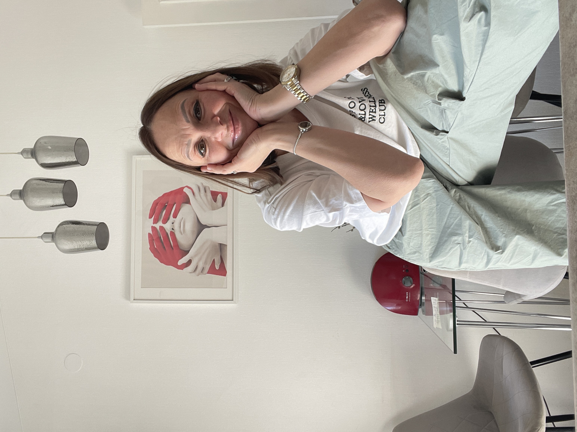 A fair-skinned woman with brown hair sits at a table. She is wearing a white T-shirt with black lettering and is resting her face in her hands. She is wearing a gold watch on her left wrist and a silver bracelet on her right wrist. Behind her is a white wall with a framed print of a person's face being covered by red hands. There are three silver pendant lights hanging from the ceiling. Also visible is a red appliance on a glass table, and a grey chair. The location is potentially a home interior. The image does not contain enough information to determine if the location is in Krasnoyarsk, Russia.