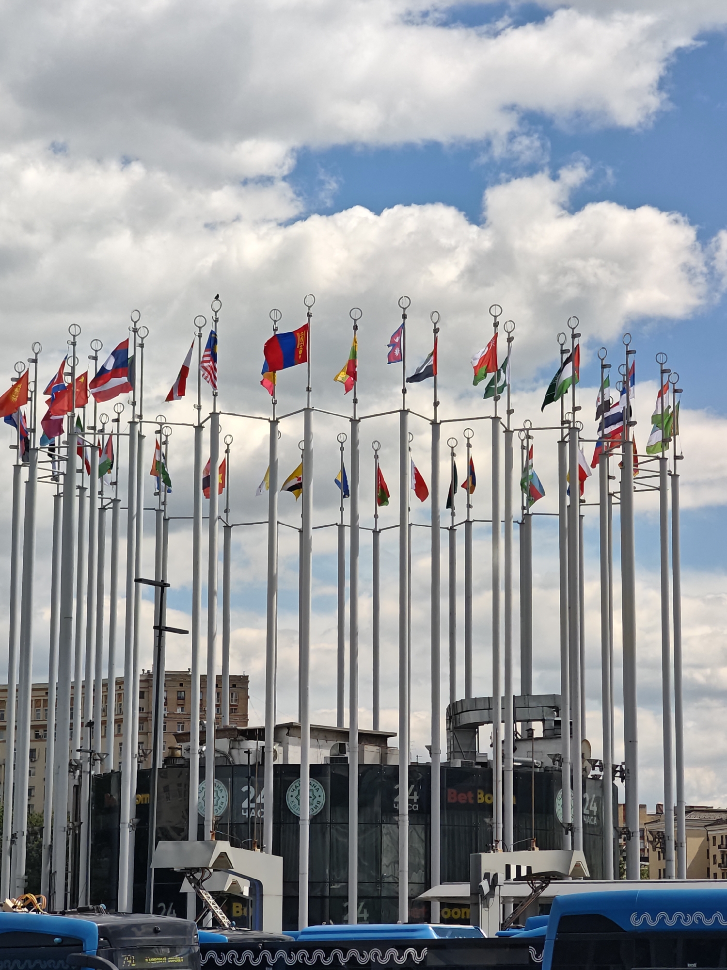 The image depicts a gathering of national flags displayed on tall poles, arranged in a circular formation against a cloudy sky. The flags represent various countries, including those of Mongolia, Malaysia, Laos, Nepal, Oman, and others that are partially obscured or difficult to identify precisely. A bird is perched atop one of the flagpoles.
In the background, a modern building with large glass windows and visible signage is partially visible, interspersed with a row of tall, thin, white poles, some of which support the flags. The building's facade features logos and text, including "BetBoom" and "24 ЧАСА" (24 Hours in Russian). Adjacent to this building, a brick building with multiple windows can be seen.
In the foreground, the roofs of several blue buses are visible, suggesting an urban or public transportation setting. The time of day appears to be daytime, with the sunlight filtering through the clouds, creating a bright but slightly overcast atmosphere. The scene is static, with the flags gently waving in the breeze. The overall impression is one of a public space or venue, possibly an exhibition area or a cultural landmark, showcasing international representation. The presence of the buses and the commercial signage on the building hints at a busy urban environment in Moscow, Russia.