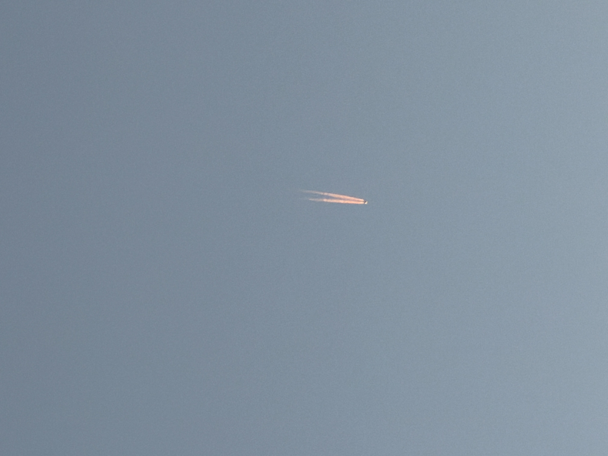 A long, straight contrail from an airplane is faintly visible in the center of the frame against a hazy blue sky. The plane is a small point at the right end of the contrail.