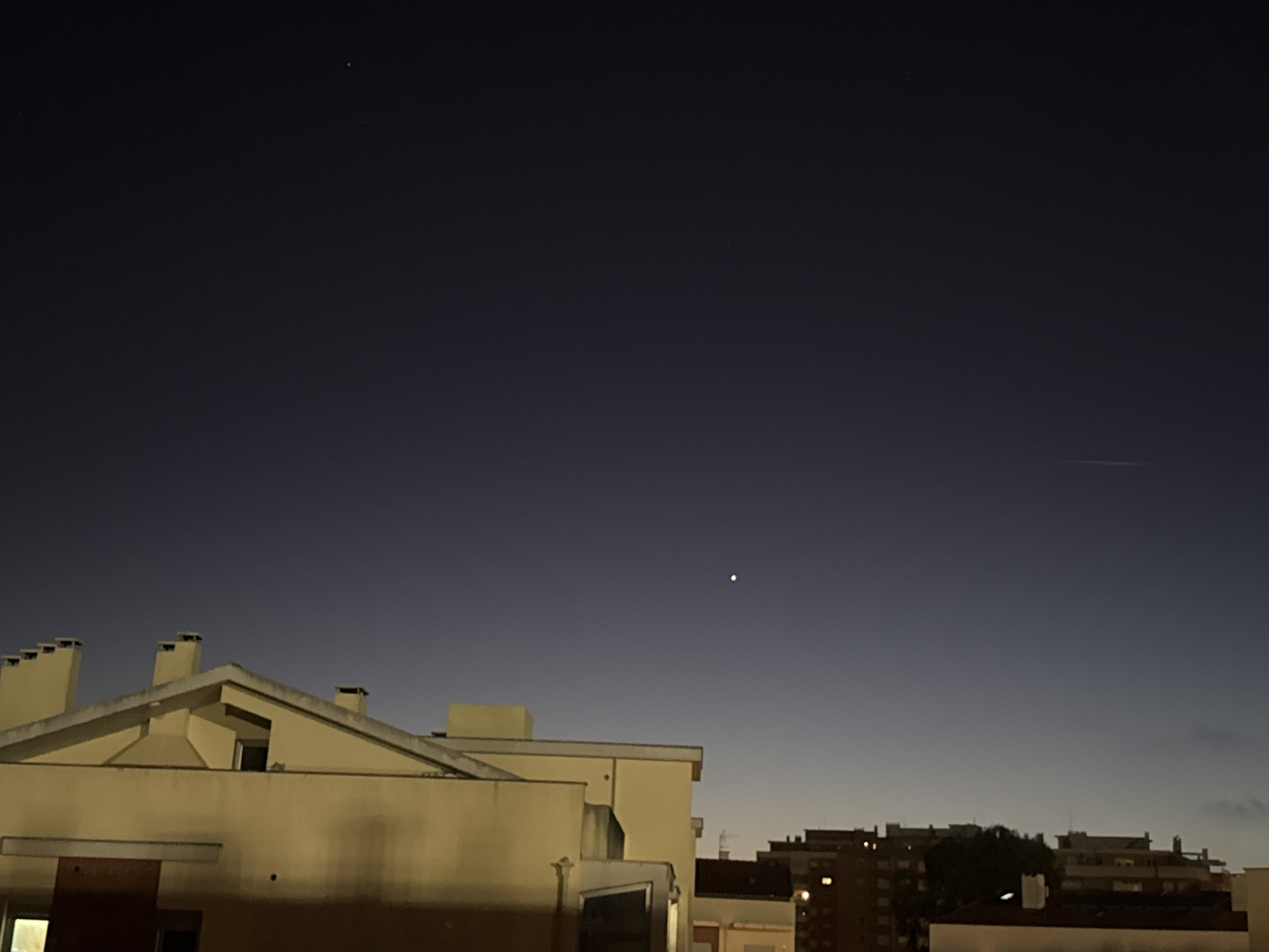 The image depicts a night scene in Figueira da Foz, Portugal. The sky is a dark blue gradient, transitioning to a lighter shade near the horizon. Several stars are visible, including a bright, isolated star in the middle of the frame and a fainter grouping higher up. A horizontal contrail is visible on the right side of the frame. In the foreground, the roofs of several buildings are visible, primarily tan-colored. The closest building has multiple chimneys. Distant buildings with lit windows are also visible in the lower-right portion of the image. There are no people visible in the image. The image captures a tranquil urban night landscape.