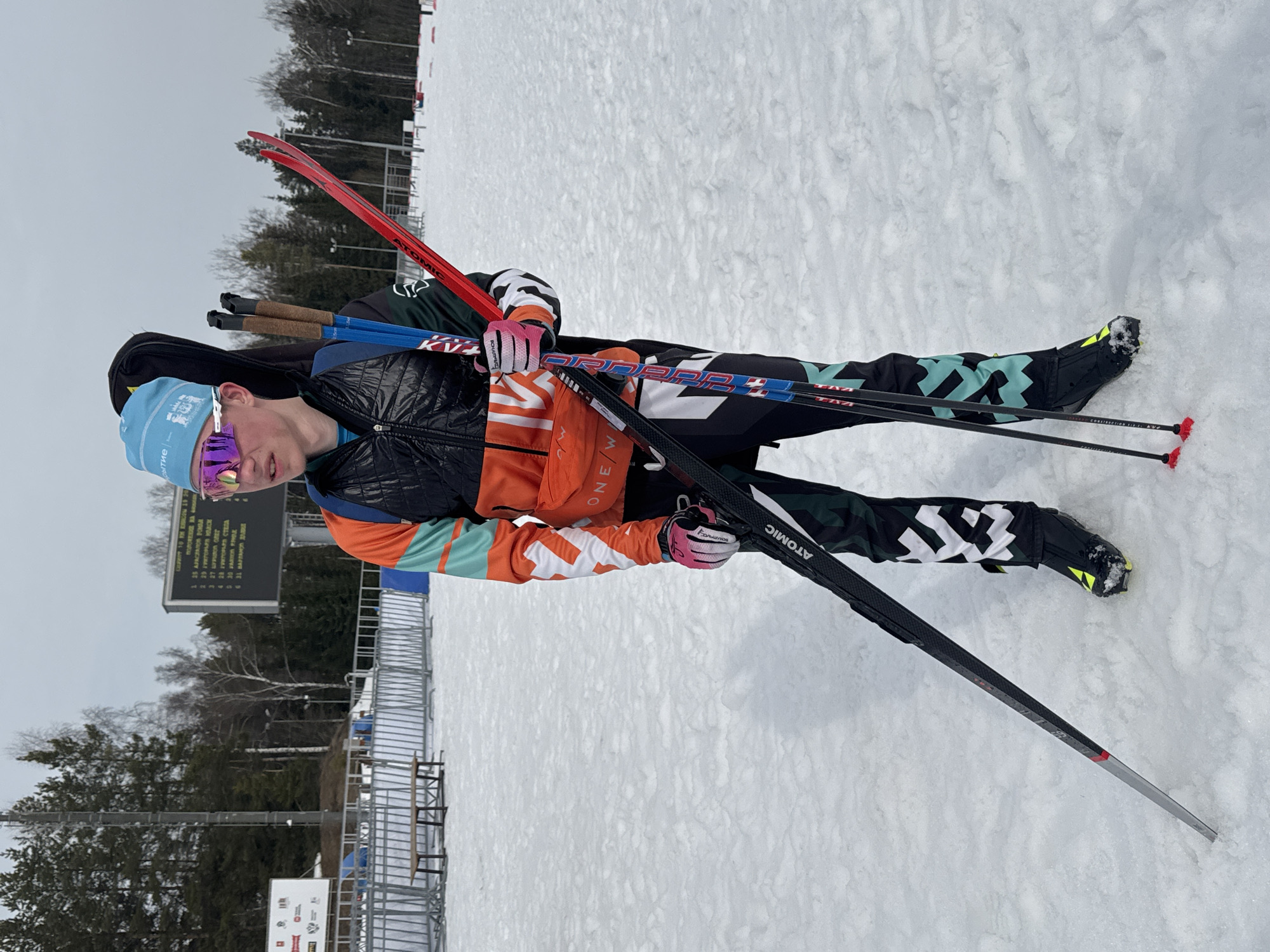 A male biathlete stands on a snow-covered ski stadium in Прикамский, Russia. He wears a light blue beanie, large purple-orange reflective ski goggles, a black padded vest over an orange and teal patterned jacket, black patterned ski pants, black ski boots with yellow accents, and pink gloves. A black ski case is visible on his back. He holds multiple sets of skis, including a pair of red skis and a pair of black 'ATOMIC' branded skis, along with two pairs of ski poles featuring blue/black shafts and red tips. The subject faces left, looking off-frame. In the mid-ground, a large digital scoreboard displays Cyrillic text, likely race information or results. Metal barrier fences and a wooden bench are present behind the subject. The distant background consists of a dense forest line with a mix of evergreen and bare deciduous trees under an overcast grey sky. The ground is uniformly covered in packed snow.