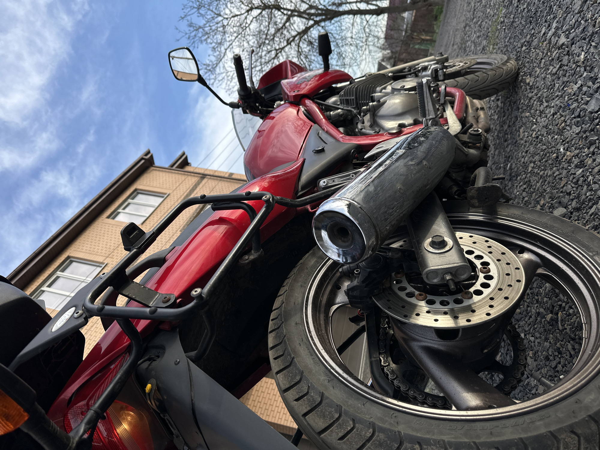The image shows a red motorcycle parked on a gravel surface. The motorcycle is positioned at an angle, with the front wheel and exhaust pipe prominently displayed. A building is visible in the background, constructed of light brown brick with white-framed windows. The sky is blue with scattered clouds. The location is potentially Rostov-on-Don, Russia, although there are no immediate identifiers in the image to confirm this.