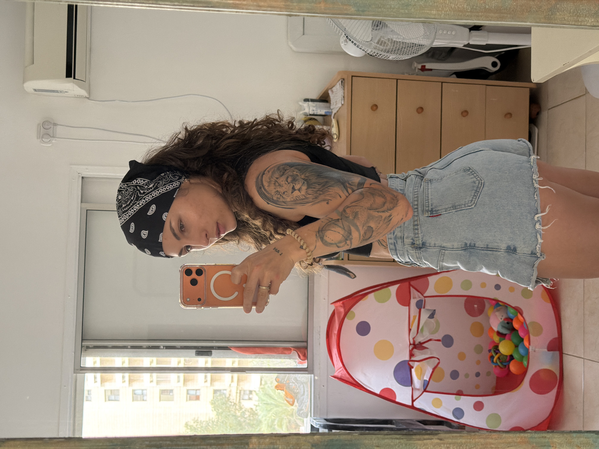 A woman with curly brown hair and multiple tattoos, including a large lion on her left arm, stands in front of a mirror taking a selfie with an orange iPhone. She is wearing a black bandana, a black top, and denim shorts.
Behind her, a dresser with drawers and a white fan on top are visible. Next to the woman is a child's play tent filled with colorful balls. A window behind the tent shows a view of a building with multiple stories, possibly in Kiryat Bialik, Occupied Palestine.
