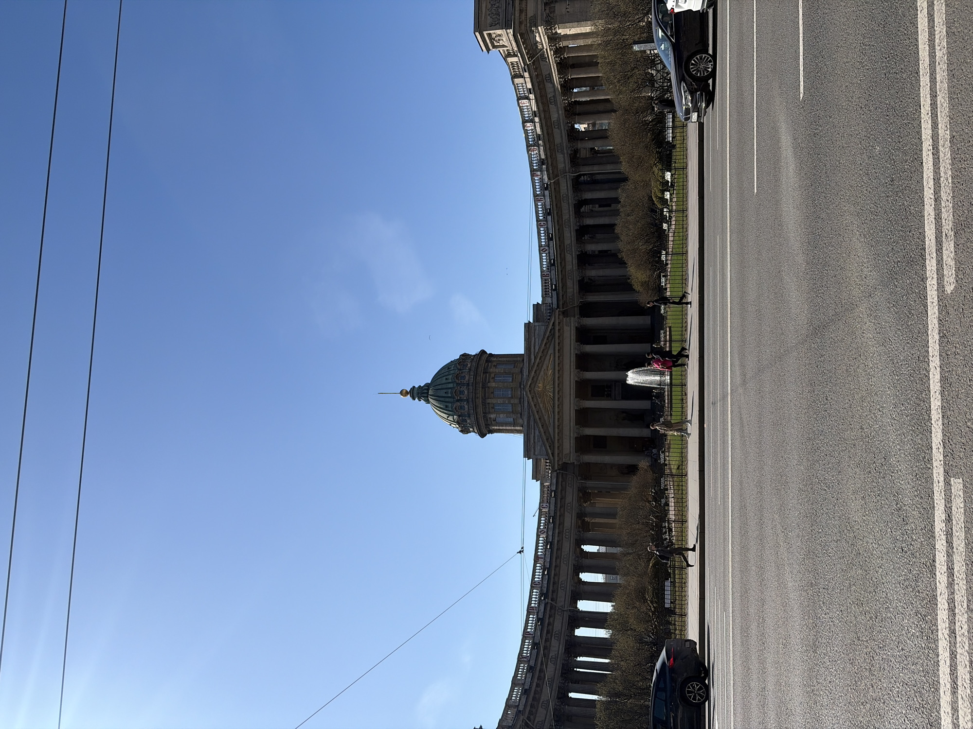 This image captures the Kazan Cathedral in Saint Petersburg, Russia, under a clear, bright blue sky. The grand Neoclassical building, characterized by its prominent green dome topped with a spire and its extensive curved colonnade, dominates the left side of the frame.
In the foreground, a wide asphalt road with white lane markings stretches across the image. A black car is visible on the right, parked along the roadside, with another vehicle partially seen further down the street. Separating the road from the cathedral is a paved sidewalk, a strip of green space with hedges or small trees, and a black decorative fence.
Several people are scattered along the sidewalk and near the cathedral, some walking, others standing. A fountain in front of the cathedral is visibly active, spraying water. The bright, uniform light indicates it is daytime, likely late morning or early afternoon. The weather appears sunny and pleasant. Overhead wires, possibly for trams or trolleybuses, are visible stretching across the upper left portion of the clear sky. No discernible text is visible in the scene.