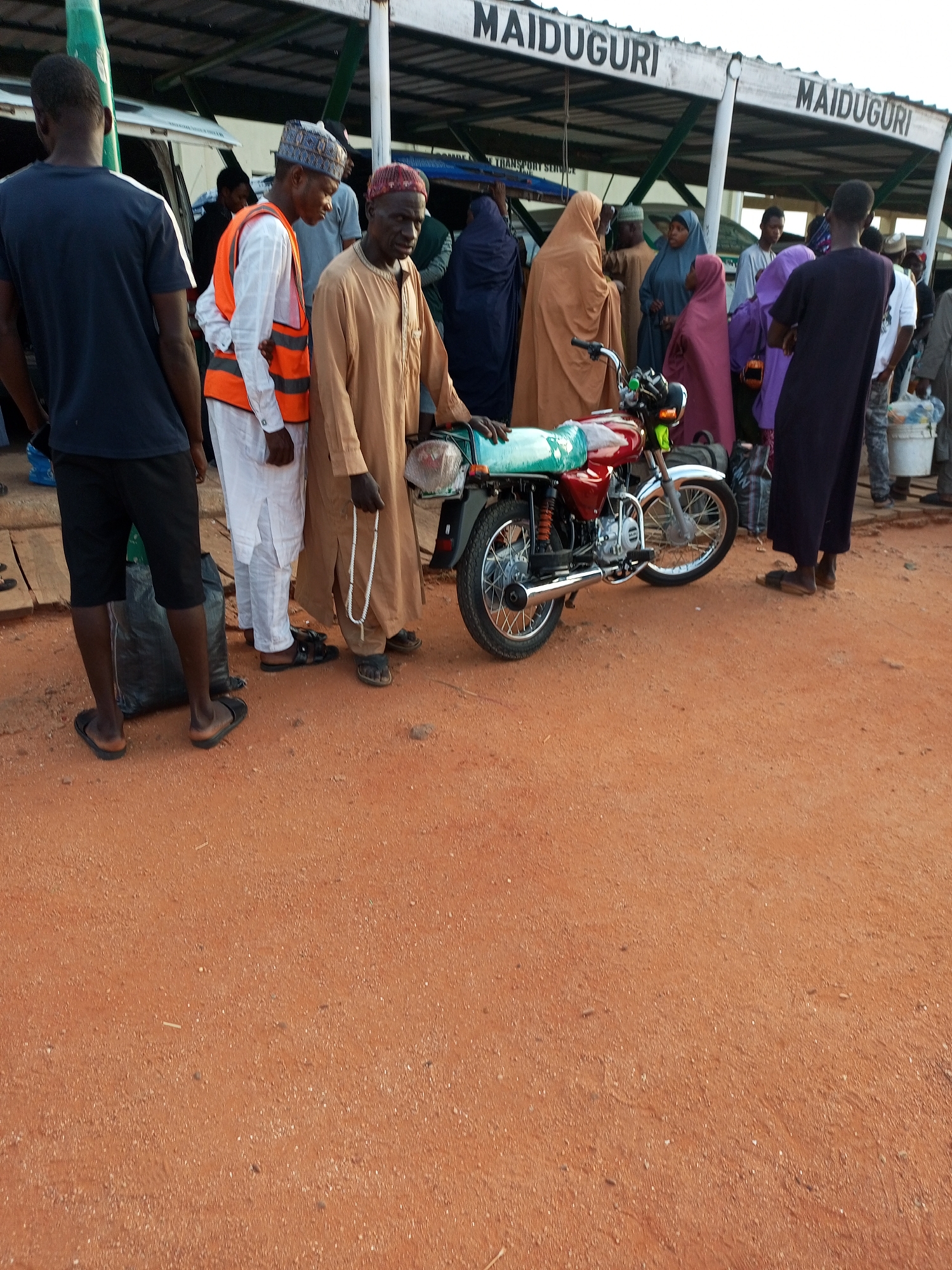 A crowd of people are gathered at a bus station in Maiduguri, Nigeria, near a red motorcycle. The ground is covered in red dirt, typical of the region. The people are dressed in a mix of traditional and modern clothing, with some wearing hijabs and others in t-shirts and shorts. Some are standing and talking, while others appear to be waiting for transportation. A bus or large vehicle is partially visible in the background. The scene appears to be a typical day at a busy transportation hub.