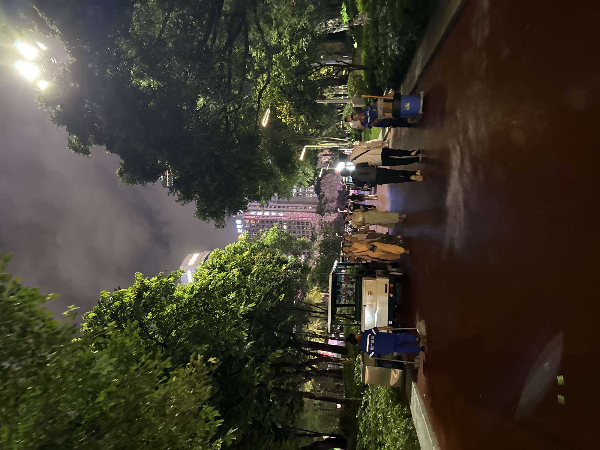 A wide, dark red paved pathway runs diagonally through a park-like setting at night, bordered by lush green trees and manicured bushes. In the background, illuminated by streetlights and the glow of distant city buildings, a group of people walk along the pathway. A white golf cart is parked on the left side of the path, with a person in a blue uniform sweeping the ground in front of it. Further down the path, two individuals, one in black and one in white, walk away from the camera. To the right, another person in a blue uniform pushes a cleaning cart. The sky above is dark and cloudy, with bright streetlights casting a yellow-white glow through the trees. The scene appears to be in Tianhe District, China, as indicated by the presence of tall modern buildings in the distance.