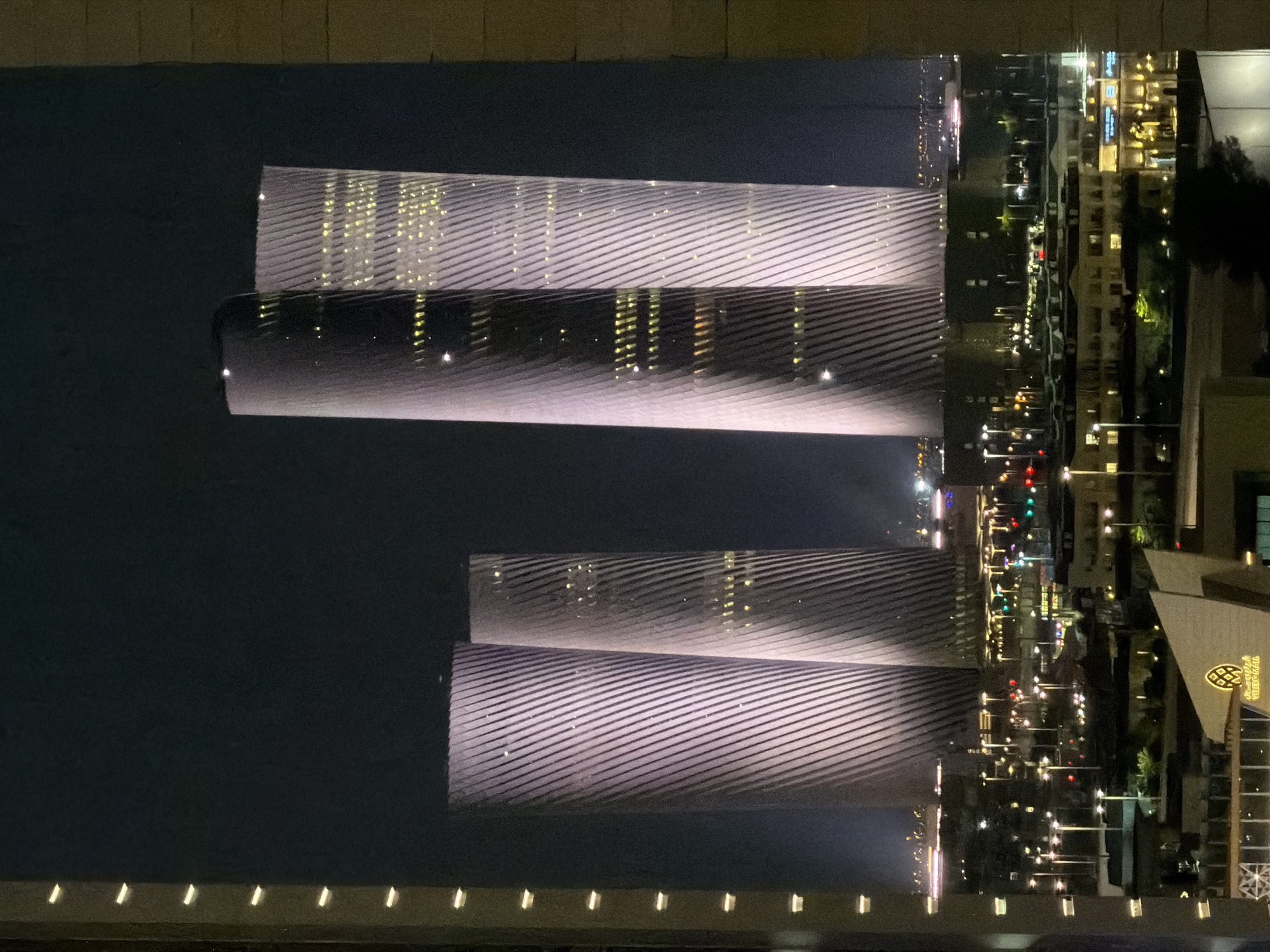 The image shows a nighttime view of the Doha skyline, featuring the iconic Tornado Towers, which are illuminated with a purple light and feature a distinctive diagonal pattern. The towers are prominently positioned in the center of the frame, with several other illuminated buildings and a cityscape visible in the foreground and background. The sky is dark, indicating nighttime, and the overall scene is lit by the artificial lights of the city. The image is framed by what appears to be a window or balcony railing on the left and right sides.