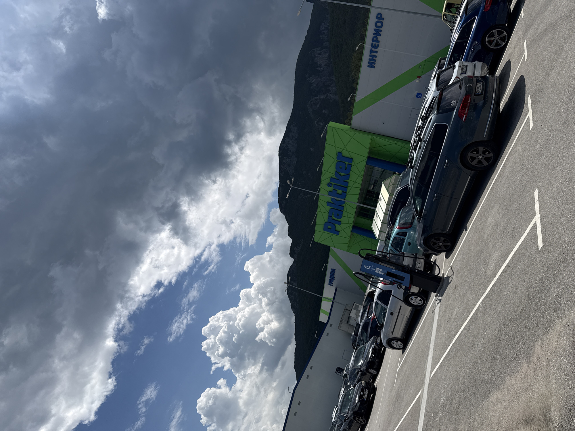 This image depicts a parking lot in front of a retail complex in Vratsa, Bulgaria, under a sky with contrasting weather conditions. The primary structure is a large commercial building, part of which is identified by the prominent green "Praktiker" logo with blue text. Adjacent sections of the building display "ГРАДИНА" (Garden) and "ИНТЕРИОР" (Interior) in Cyrillic, also on a white facade with green accents.
The parking lot is asphalted with white line markings delineating parking spaces. Numerous passenger vehicles are parked, oriented diagonally to the left. In the mid-ground, a blue electric vehicle charging station, bearing the "EVN" logo, stands prominently. Vehicles visible include a dark gray Volkswagen Passat station wagon with roof rails in the immediate foreground (license plate "BP 3556 CM"), a silver Opel Corsa, a black SEAT Leon (license plate "BP 6972 CA"), and a blue Audi station wagon further back.
In the background, behind the retail complex, a densely forested mountain range rises under the sky. The sky itself is dramatically divided; the upper left portion features bright blue sky with large, fluffy white cumulus clouds, while the upper right and central areas are dominated by dark, heavy, gray clouds, indicative of an impending or recent storm. The sunlight is strong on the left side of the scene, casting shadows, while the right side appears dimmer under the heavy cloud cover. No people are visible.