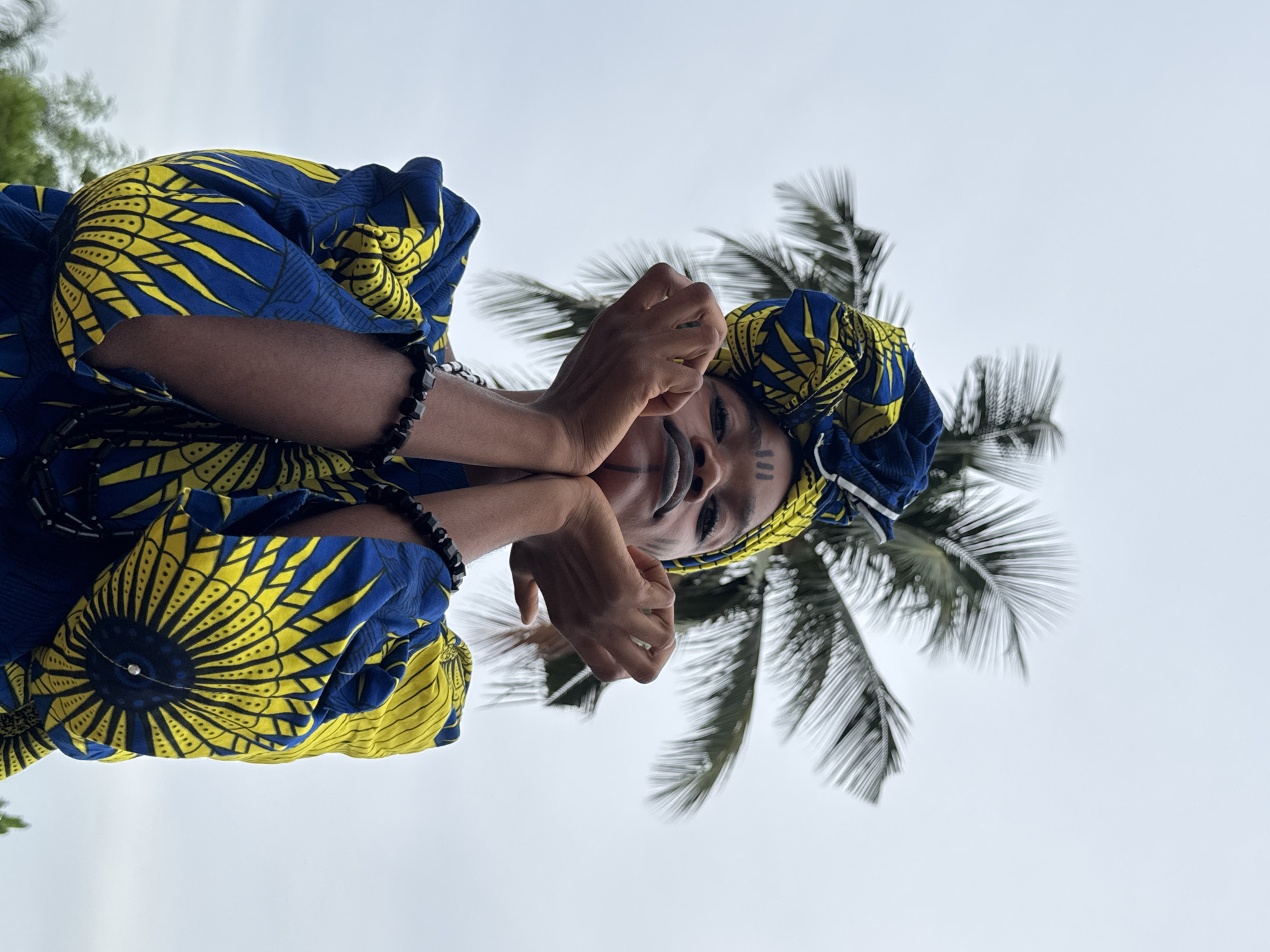 A person wearing traditional Nigerian clothing and face paint stands beneath a palm tree in Badagry, Nigeria. The person is wearing a blue and yellow patterned outfit and a headwrap of the same material. They have three vertical lines painted on their left cheek and a line on their chin. Their hands are near their face. A black beaded bracelet is visible on their left wrist. A palm tree is behind the person. The sky is overcast.