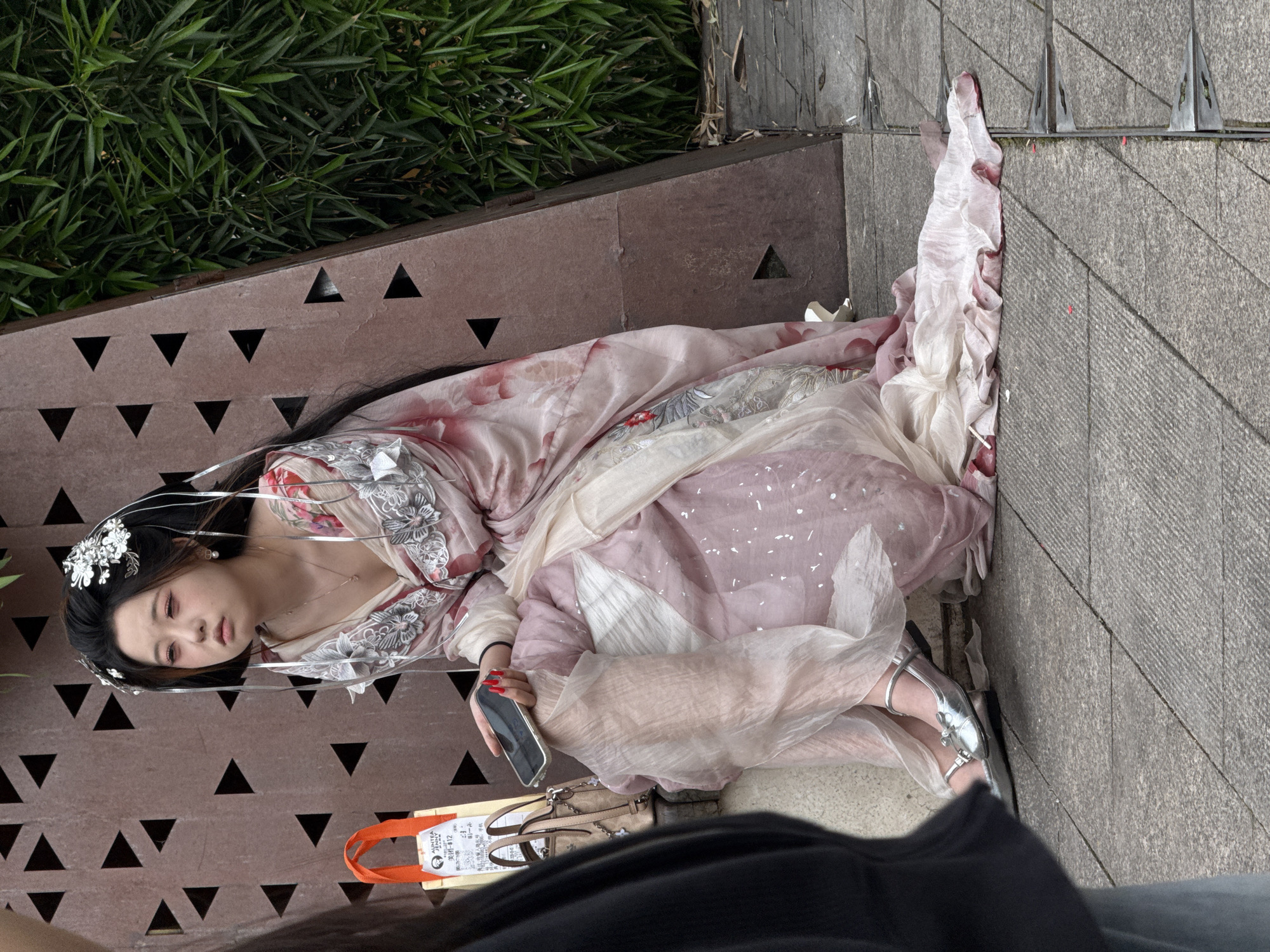 A young woman in traditional Chinese attire is reclining on an urban concrete ledge in Yuzhong District, China. She wears a pale pink and white gown, elaborate silver hair ornaments, a necklace, and silver shoes. She is holding a smartphone in her right hand and appears to be looking at it. A tan handbag rests nearby. The ledge has decorative triangular cutouts and is adjacent to green foliage. A person wearing a black shirt is partially visible in the foreground.