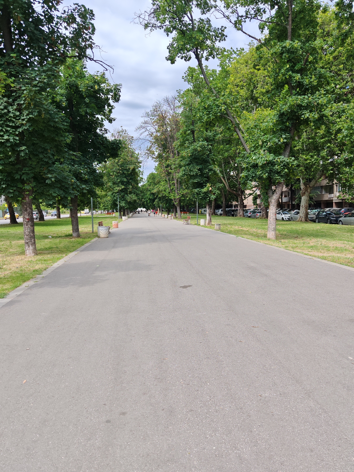 The image depicts a wide, paved walkway in a park-like setting. The walkway is flanked by mature trees with lush green foliage on both sides, creating a shaded avenue. The ground is asphalt, showing some minor wear and tear with a few scattered leaves.
To the left of the walkway, there is a grassy area with several trees and a line of parked cars visible in the distance. There are also a couple of trash receptacles and benches along the edge of the walkway.
To the right of the walkway, there is another grassy area with trees and a multi-story building in the background. More parked cars are visible in this area as well.
Further down the walkway, several people can be seen walking, suggesting it is a public space used for recreation. The sky above is overcast with some clouds.
This scene is consistent with a public park or boulevard within a city. Based on the architecture of the buildings visible and the general ambiance, this location is likely in the city of Plovdiv, Bulgaria, specifically a section of the Tsar Simeon's Garden or a similar urban green space.