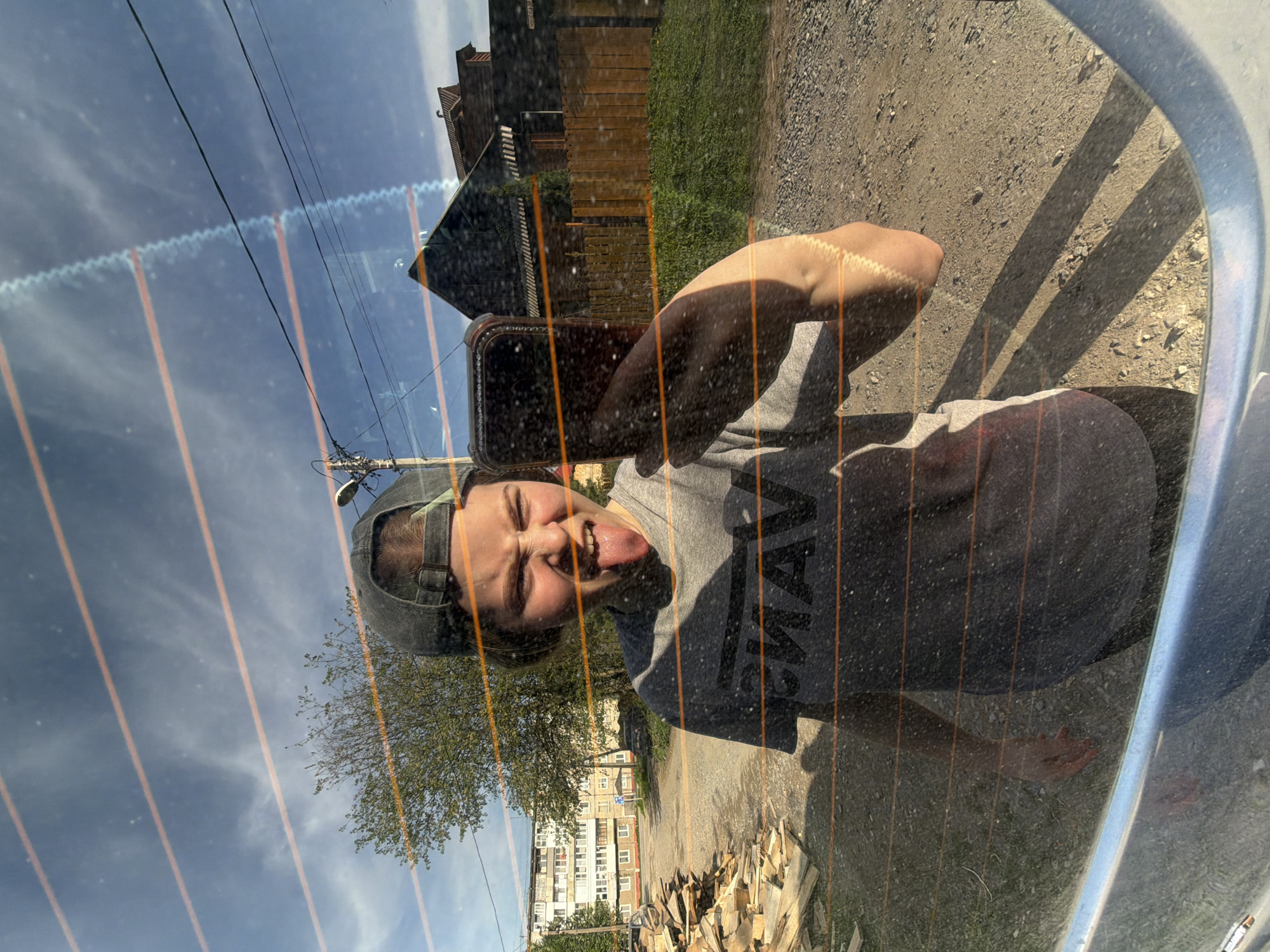The image shows a reflection in a car window, presumably taken in Perm, Russia, based on the visible buildings and style. A person wearing a baseball cap and grey t-shirt is taking a selfie with their phone, sticking their tongue out. Behind the reflection, there's a building, a tree, stacked wood, a dirt road, a wooden fence, grass, a blue sky, and some utility poles. The car window has orange defroster lines.
